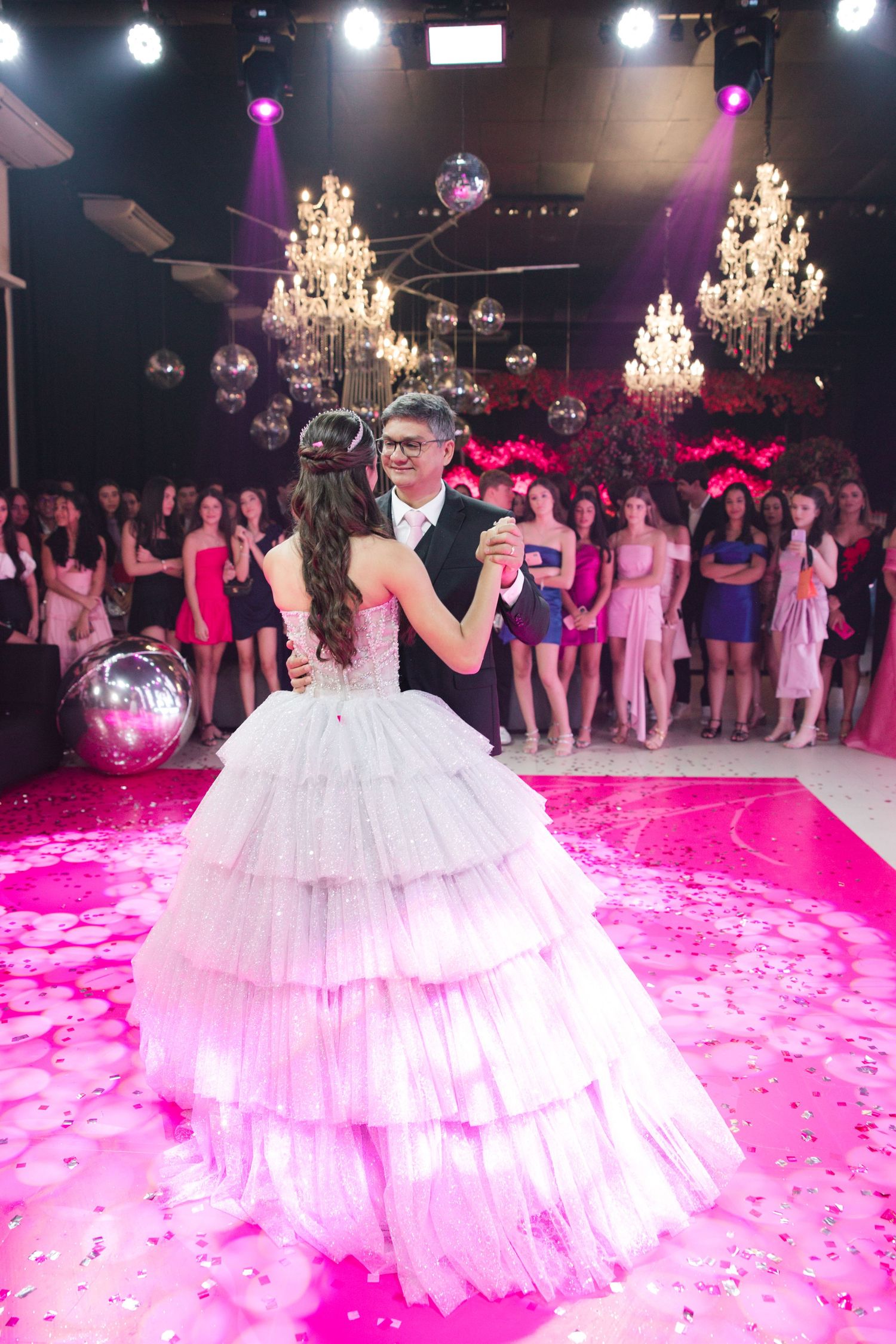 Elegant ballroom celebration with crystal chandeliers and pink lighting as couple dances surrounded by guests.