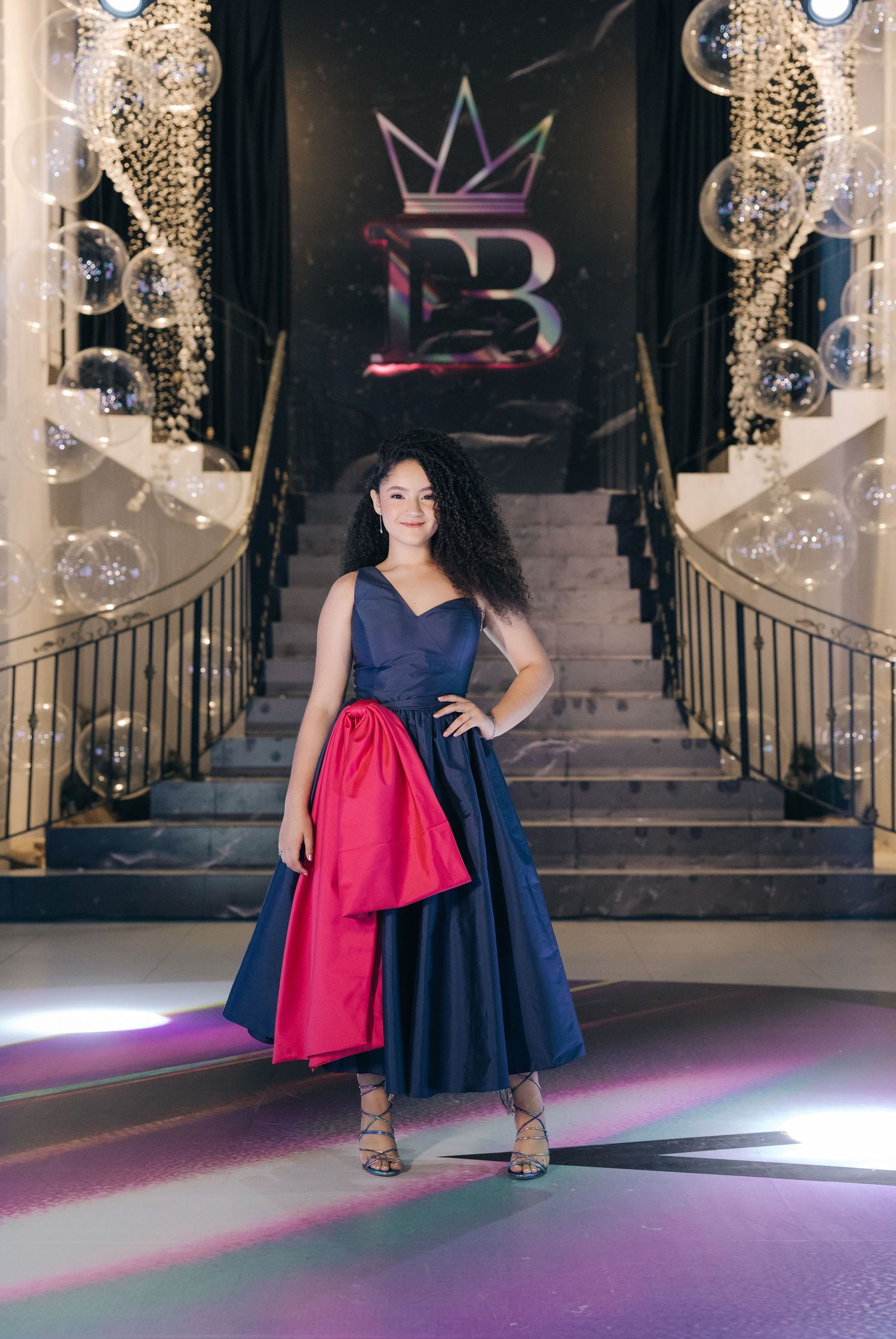 Navy and red formal dress on a grand staircase with crystal chandeliers and illuminated crown logo.