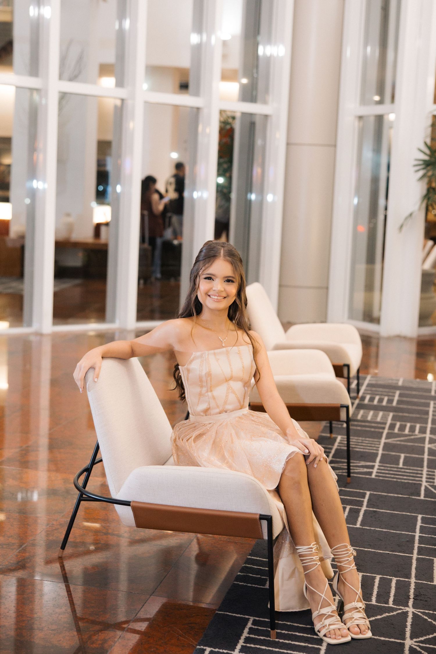 Figure sits elegantly in a modern lobby setting wearing a blush-colored dress and strappy heels against geometric floor patterns.