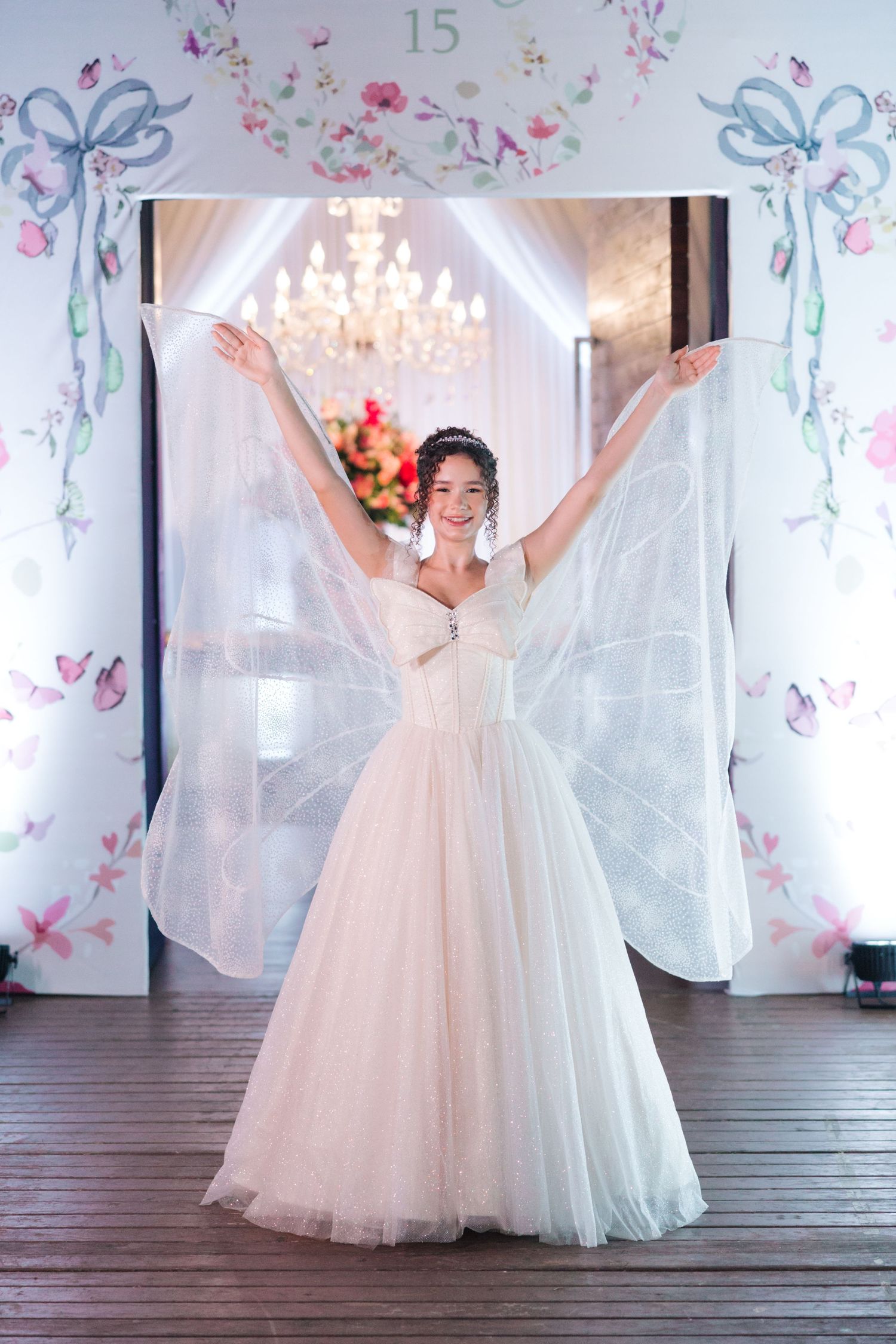 A bride in a tulle ballgown raises her arms in joy beneath a crystal chandelier and floral decorations.