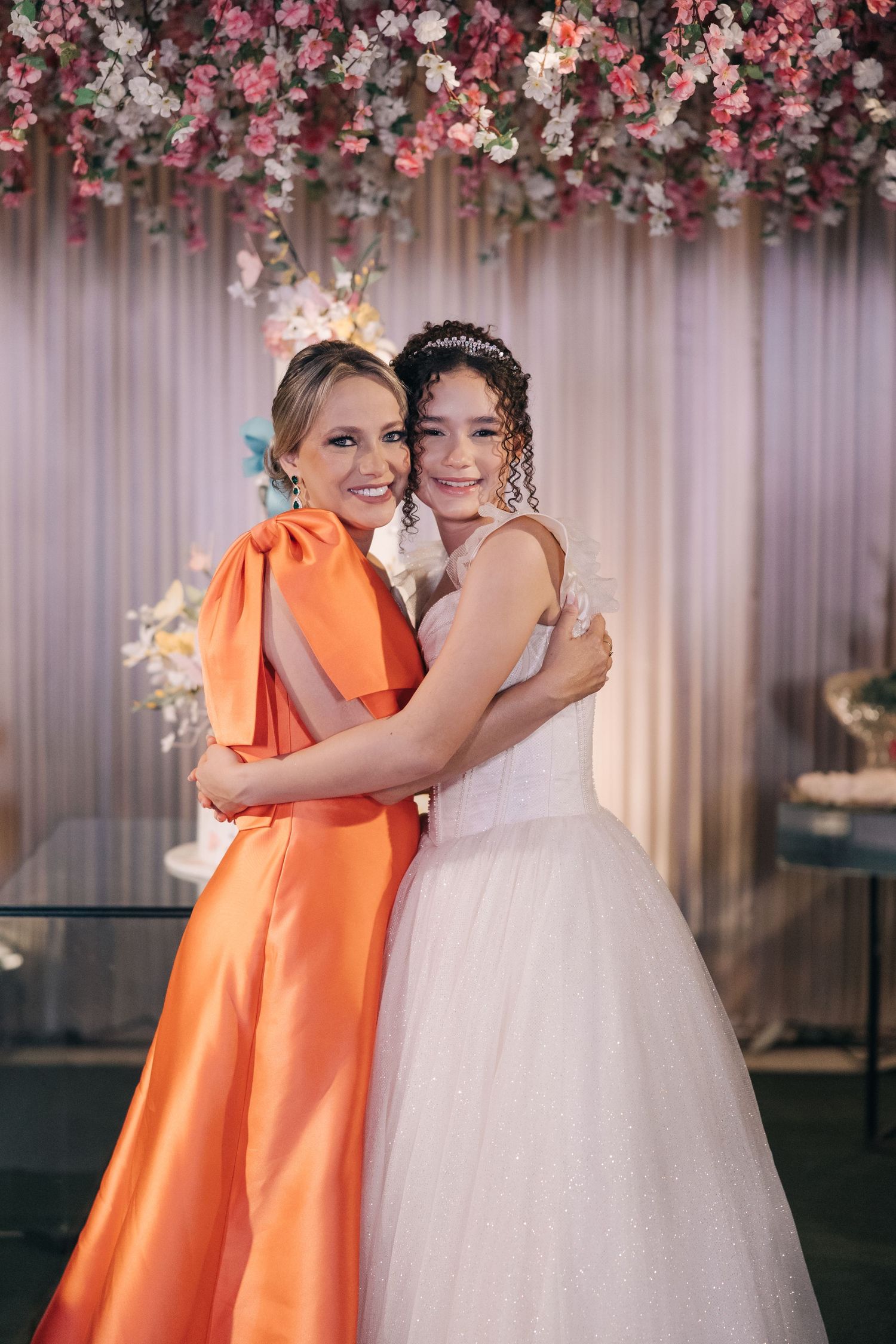 A bride in white and guest in orange pose together under a stunning floral arch display.