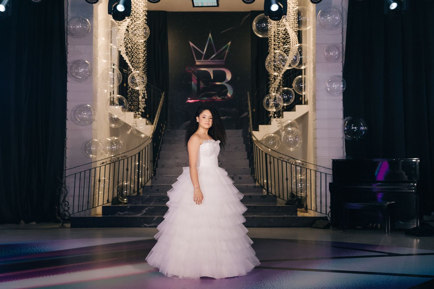 A beautiful white quinceañera dress with ruffled layers stands in an elegant ballroom entrance with illuminated decor.