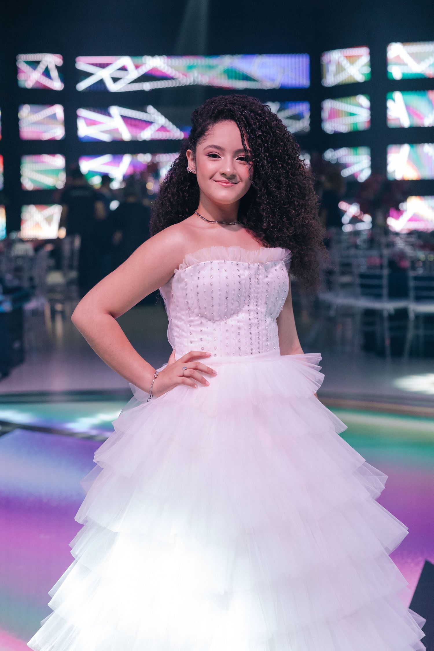 A person wearing a white layered ballgown with a fitted corset bodice poses in front of colorful digital displays.