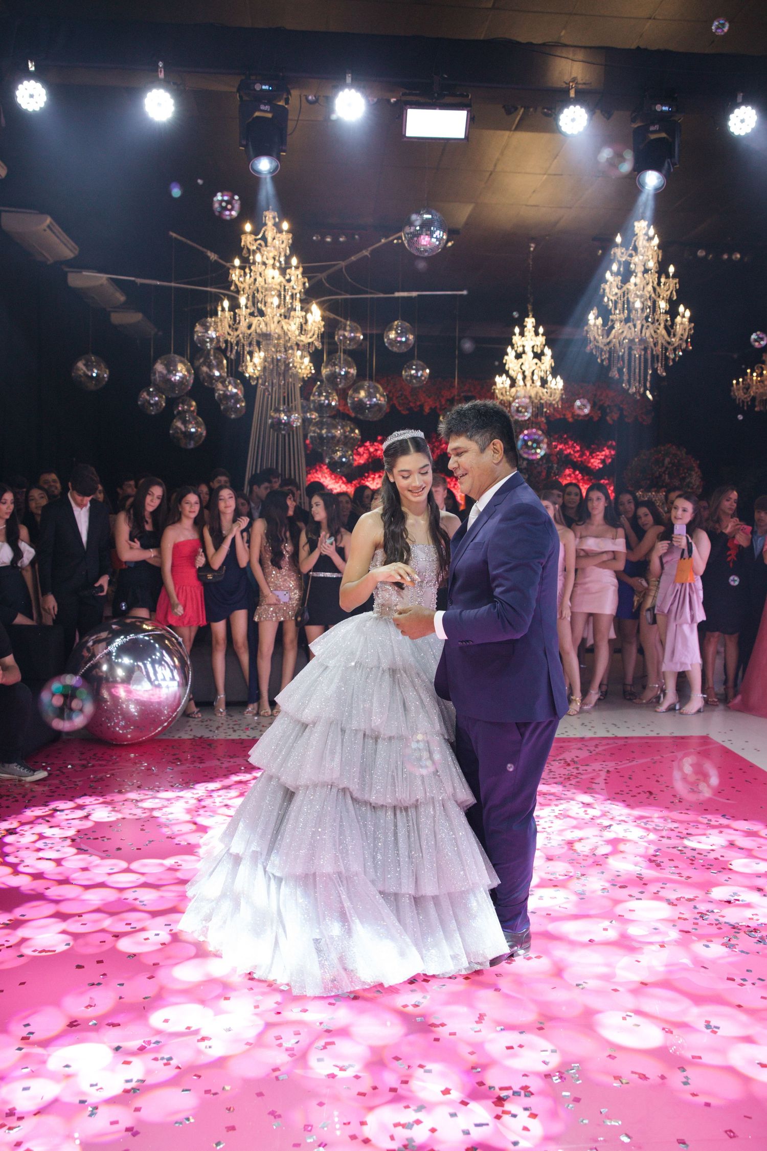 An elegant ballroom dance scene with chandeliers and pink lighting creates a romantic celebration atmosphere.