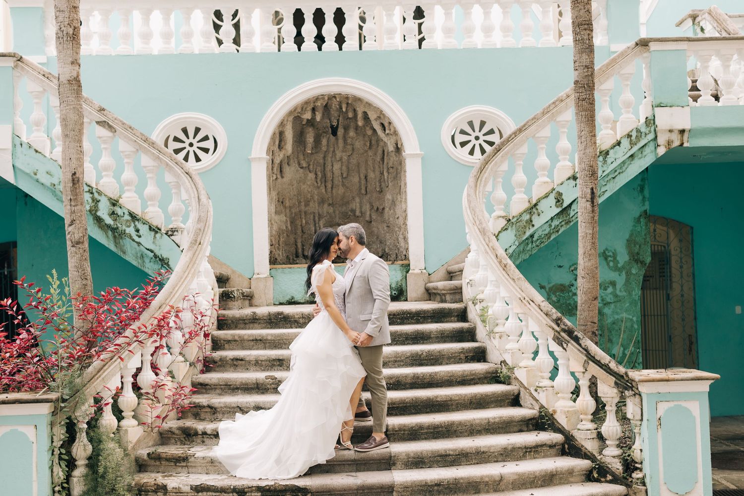 Couple embraces on a grand turquoise staircase adorned with pink flowers at an elegant colonial mansion.