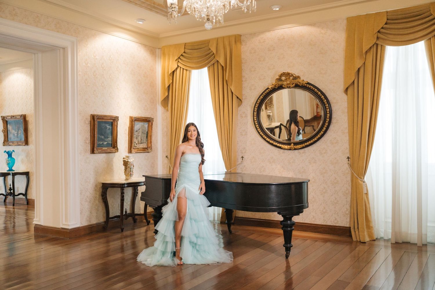 An elegant ballgown is displayed in an opulent room with golden curtains, chandelier and grand piano.