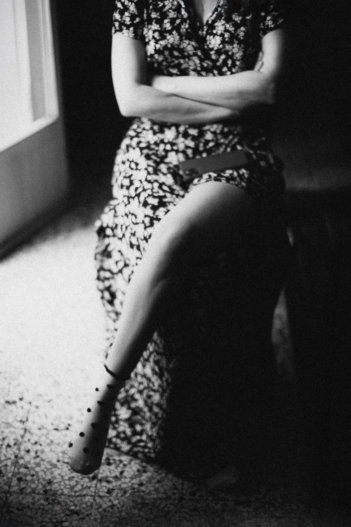 A black and white photo shows a person seated by a window wearing a floral dress with crossed arms and legs.