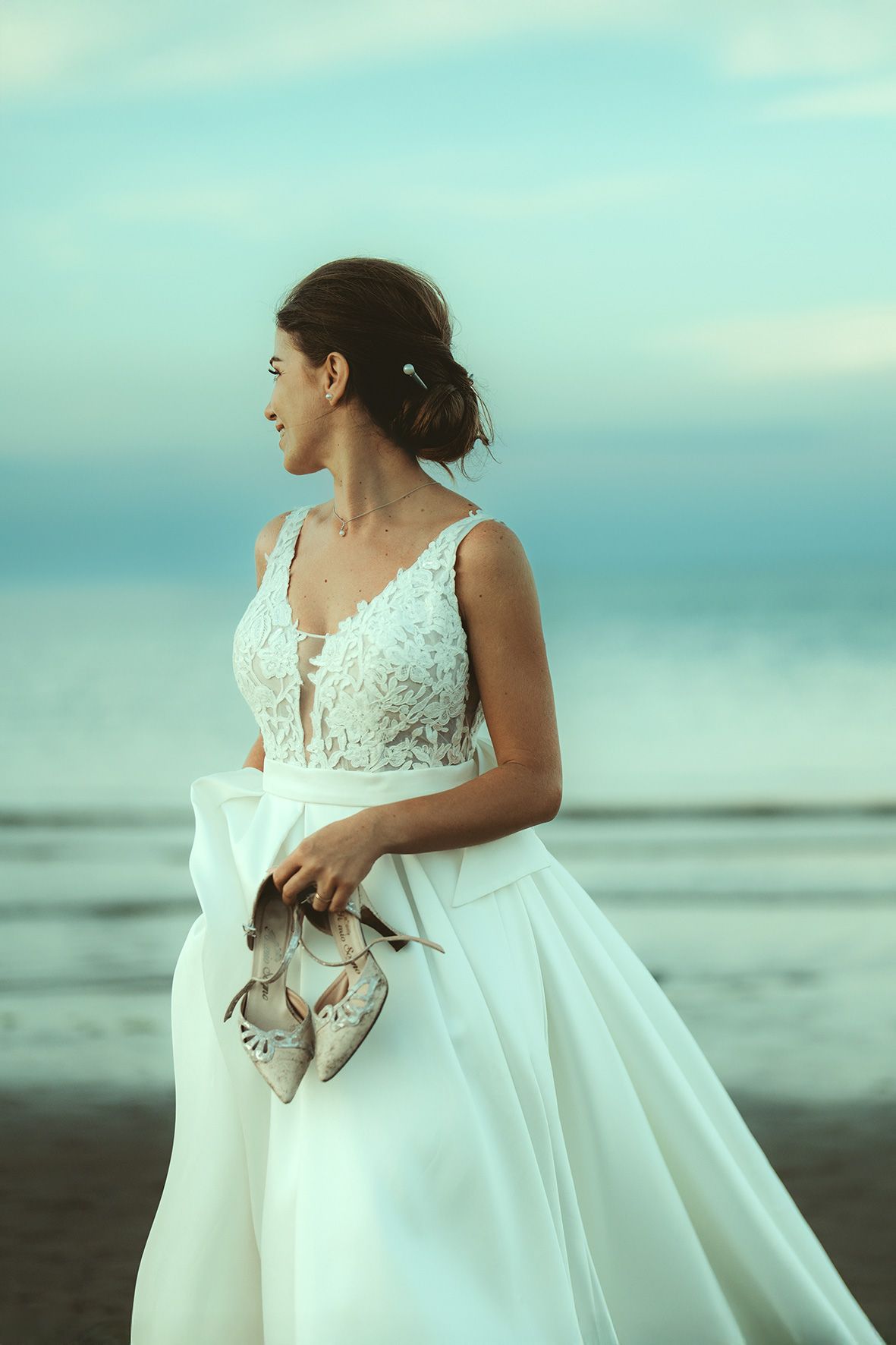 Elegant bride in a lace wedding dress holding shoes on a misty beach at sunset.