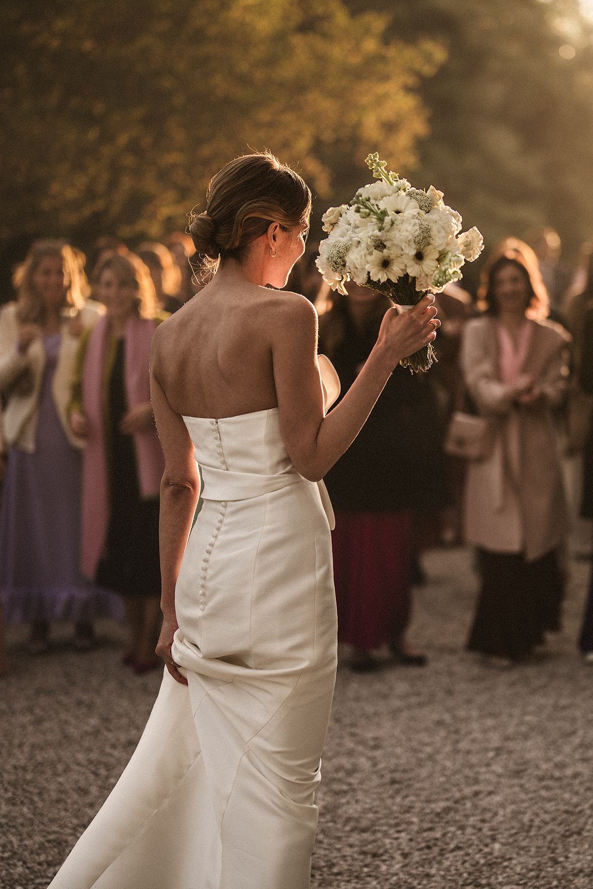 Back view of bride in classic white dress holding bouquet at outdoor sunset wedding reception with guests in background.