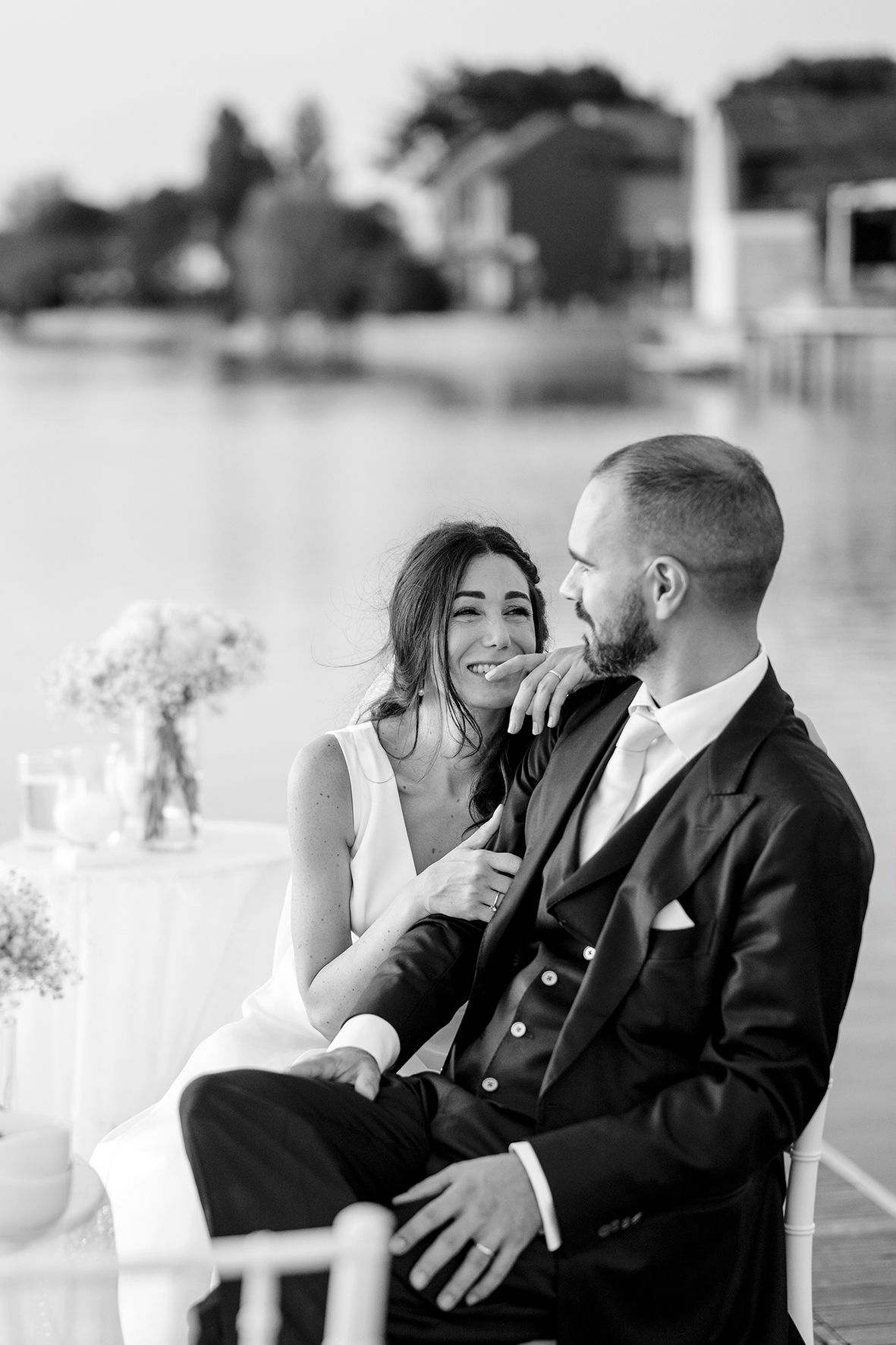 A romantic black and white wedding moment captured of a couple sharing an intimate embrace on an outdoor waterfront terrace.