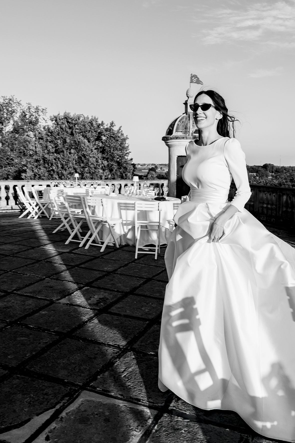 A sunlit wedding reception setup on a patio with white chairs and a person in a formal white dress.