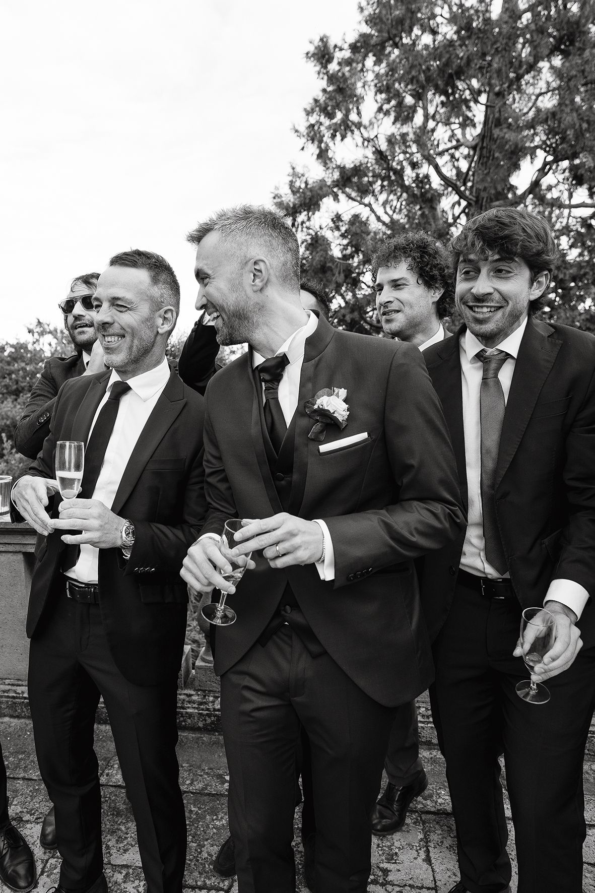 A group of men in formal black tuxedos laughing and socializing at an outdoor event.