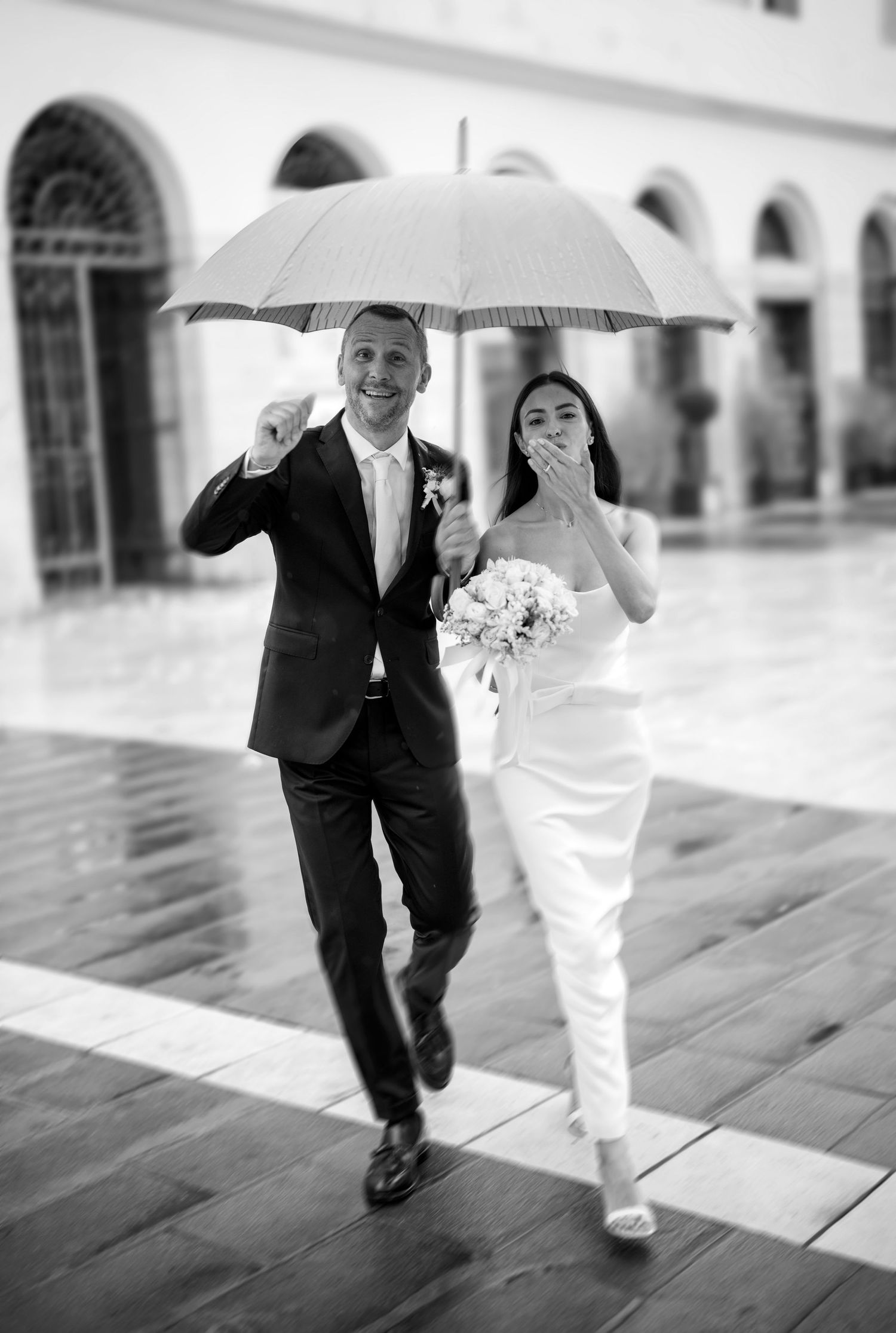 A black and white wedding photo shows a couple walking with an umbrella in an elegant courtyard with arched architecture.