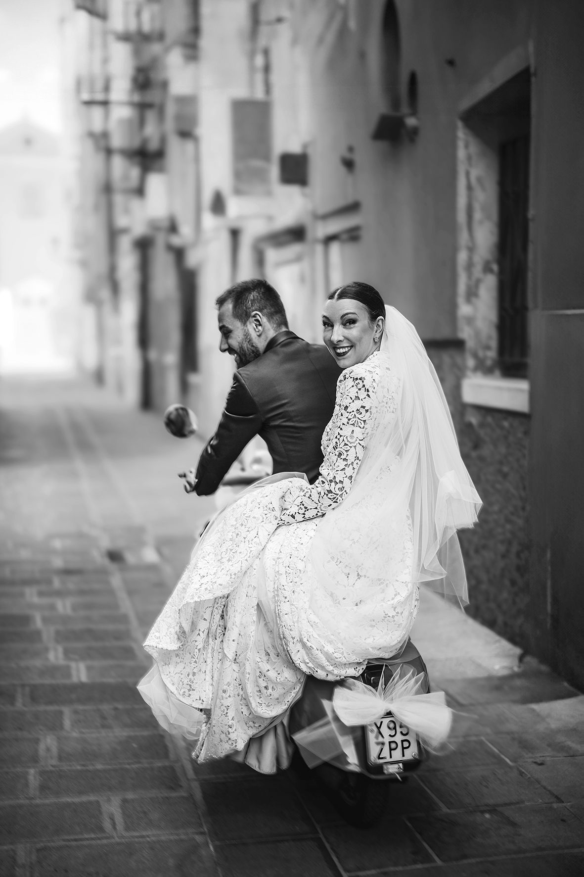 A playful black and white wedding photo shows a couple on a Vespa scooter along a charming European street.