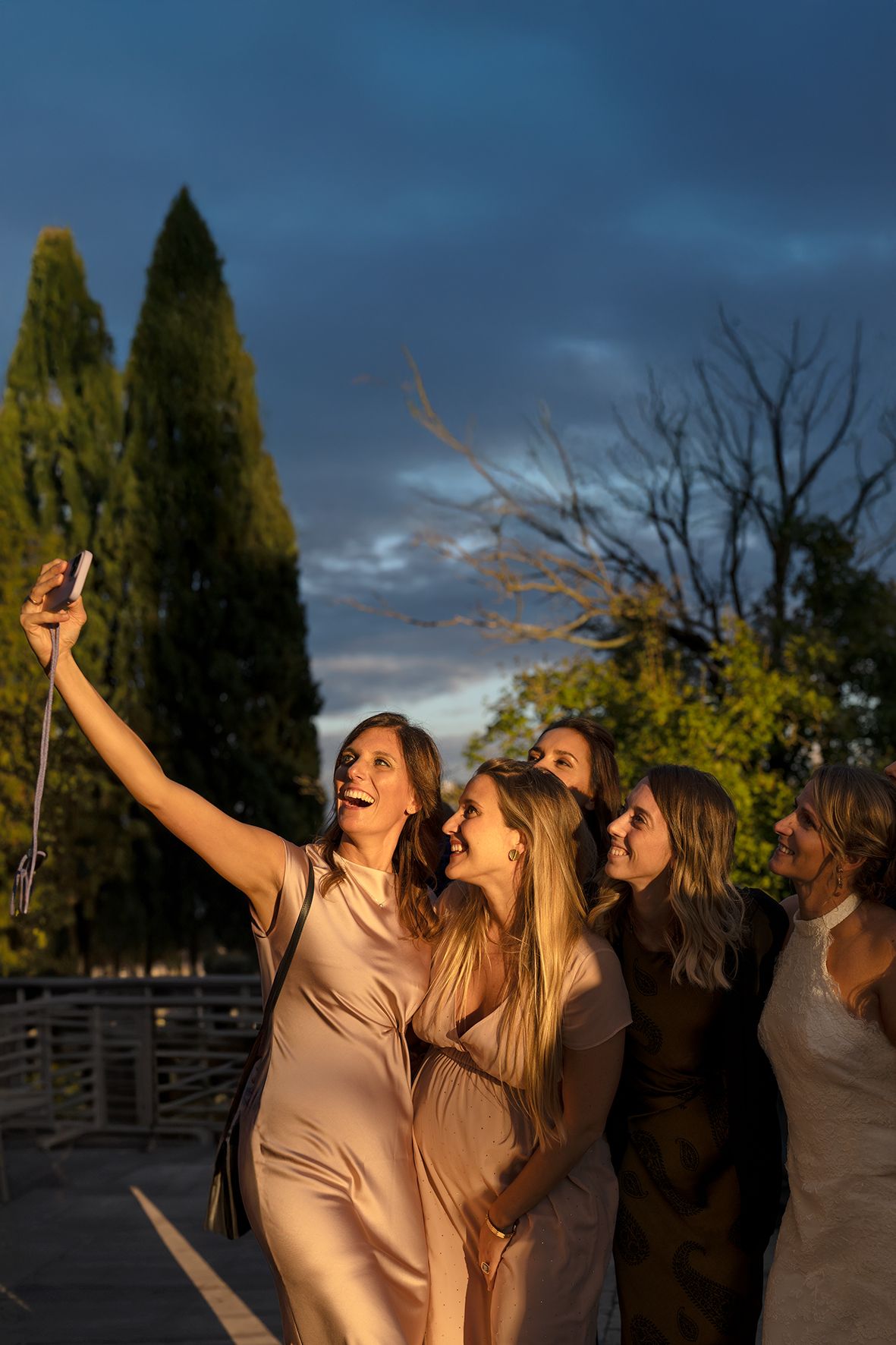 A group of friends in beige dresses laugh together while taking a selfie at sunset with cypress trees in the background.