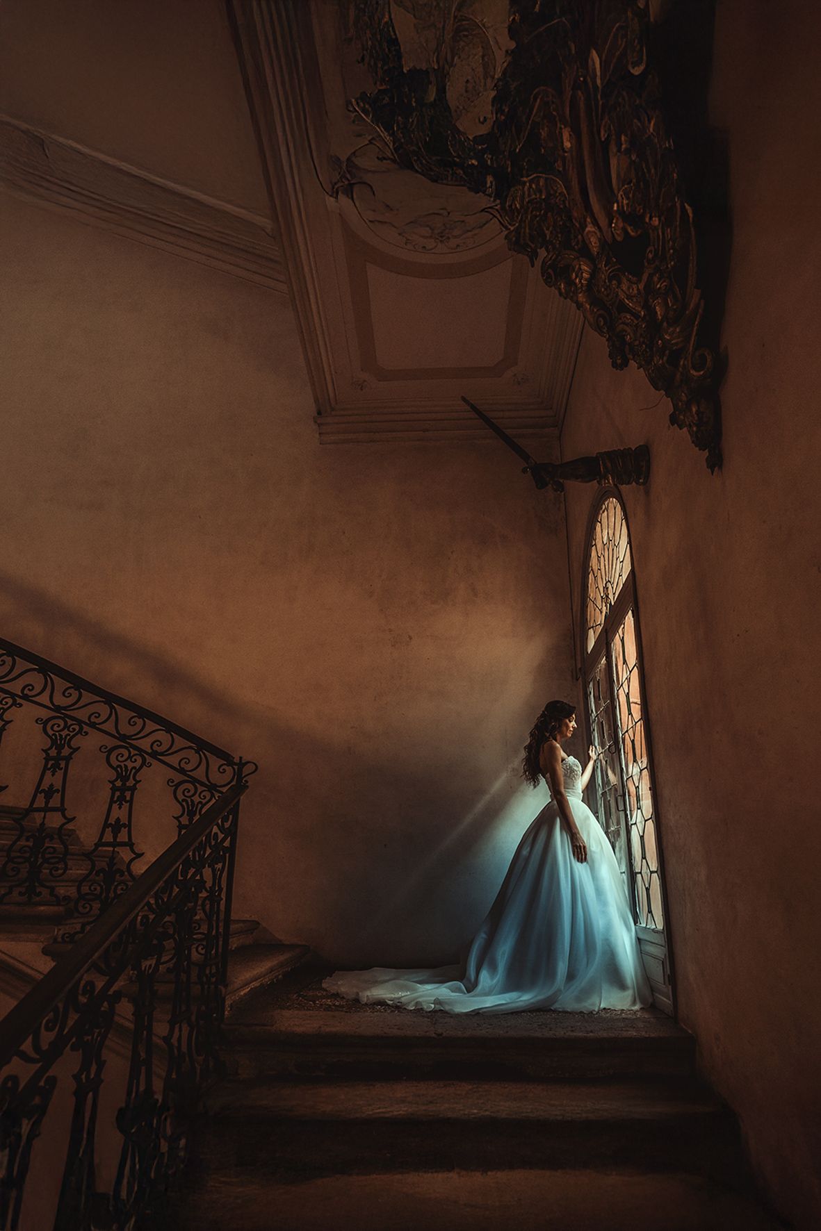 A figure in a flowing light blue ball gown stands on a dramatic spiral staircase in a dimly lit vintage interior.