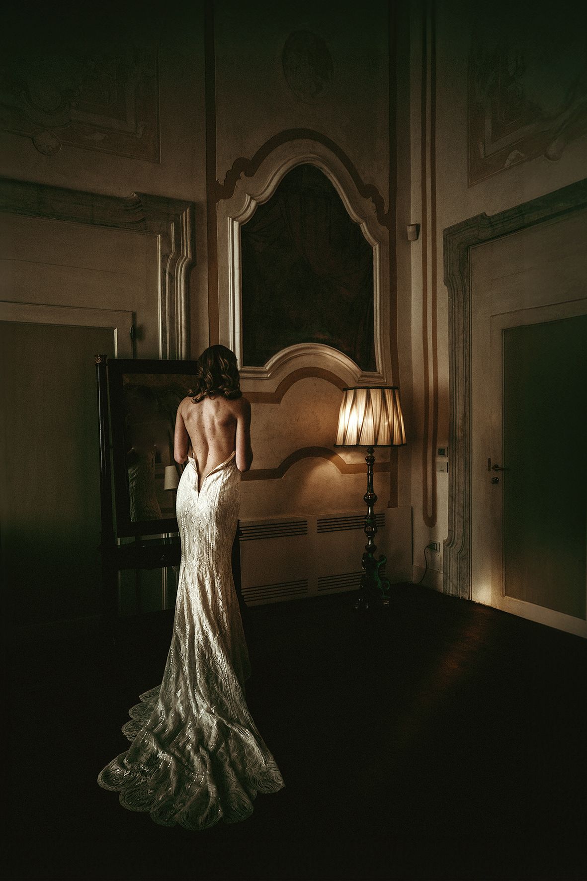 Elegant bride in backless lace gown poses in dramatic lighting against ornate mirror and lamp.
