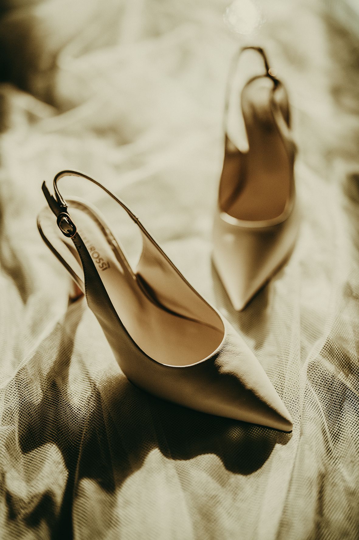 Elegant beige slingback heels with pointed toes sit on a light cream fabric background in moody lighting.