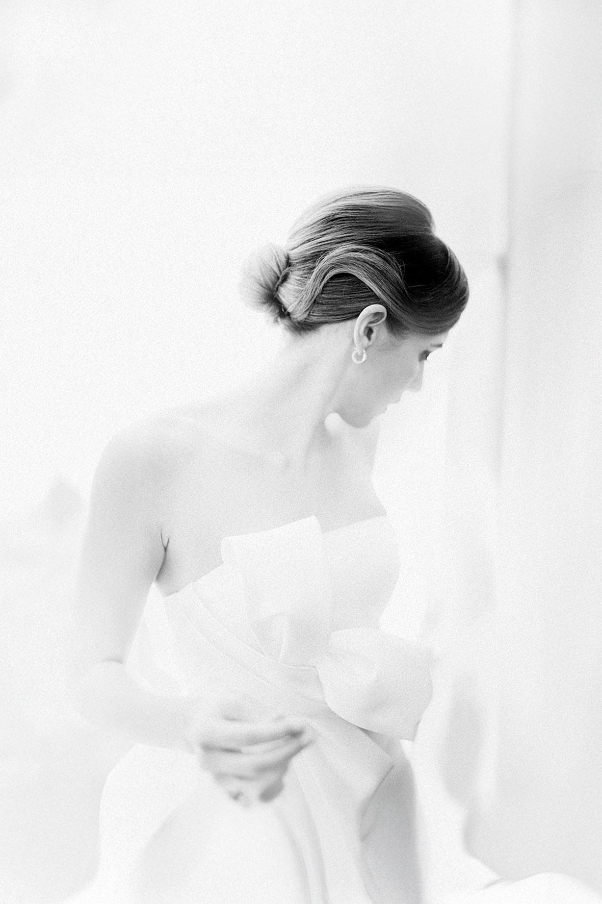 A soft black and white portrait of someone in a white strapless wedding dress gazing downward in an elegant pose.