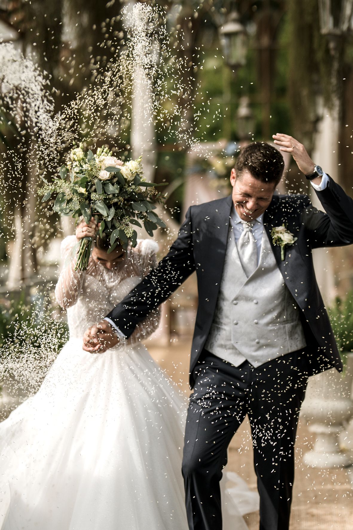 Joyful wedding couple laughs while being showered with confetti on their special day in a tree-lined setting.