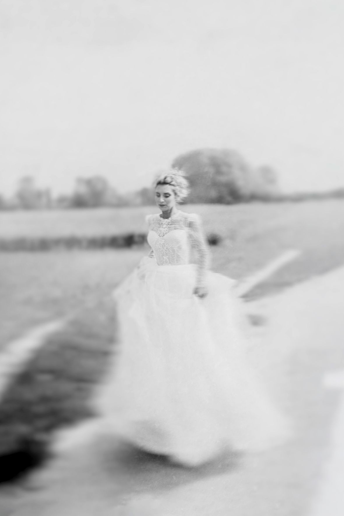 A dreamy black and white photograph of someone in a flowing white wedding dress walking outdoors in soft focus.