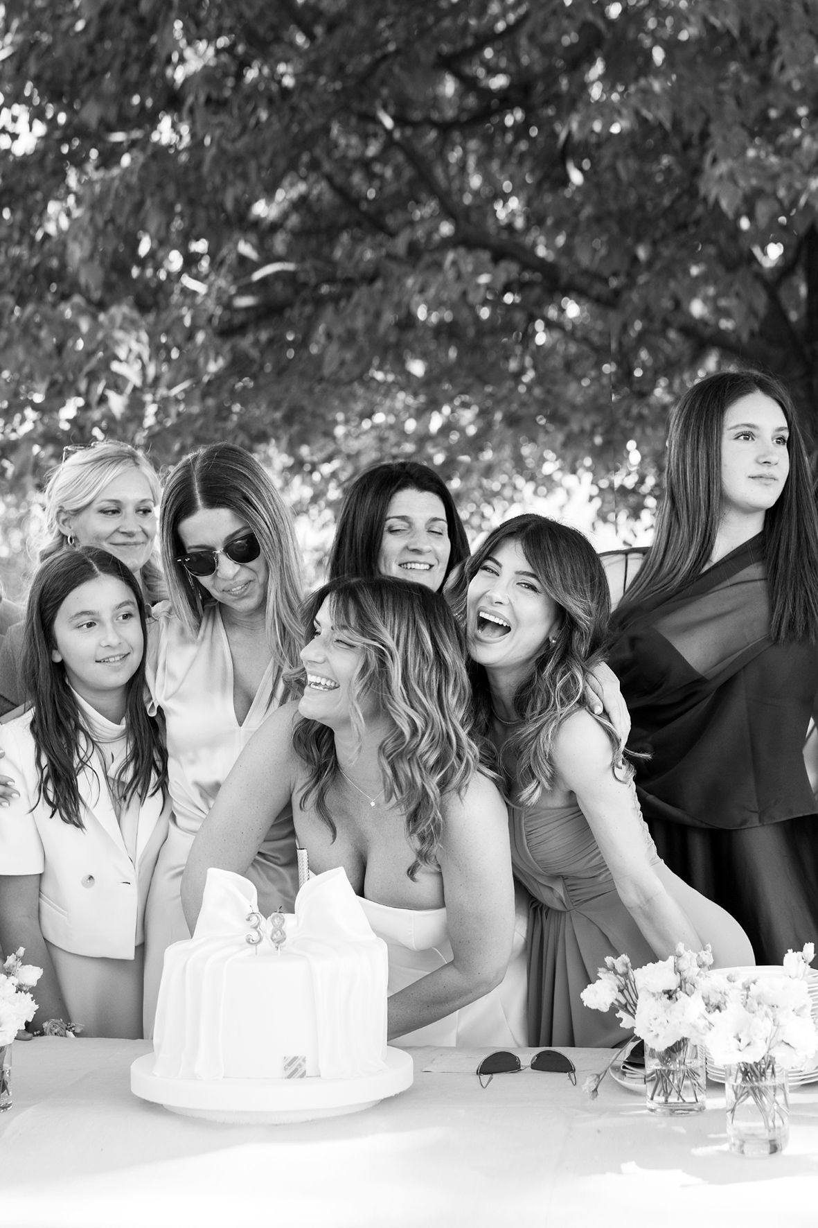 A group of women laugh together while gathered around a white cake at an outdoor celebration table.