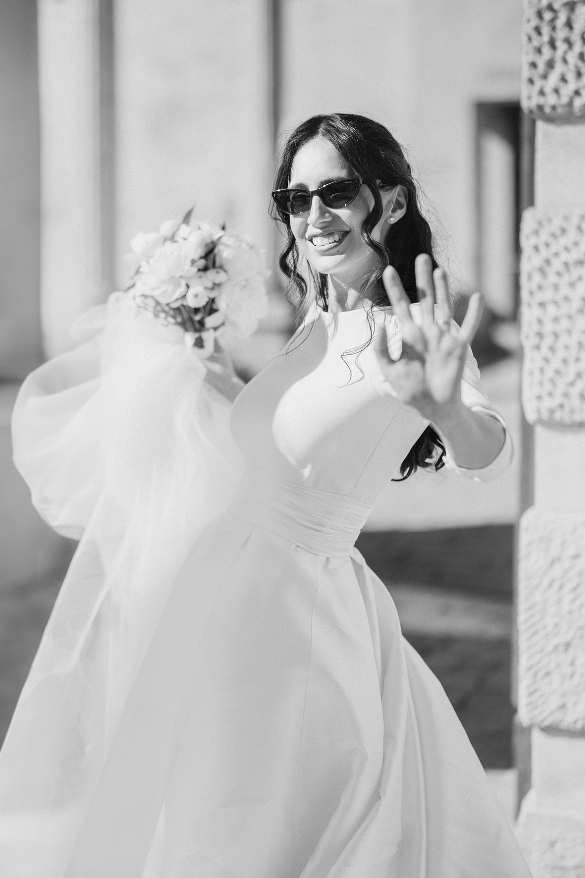 A joyful bride in a flowing white dress and sunglasses waves at the camera in black and white.