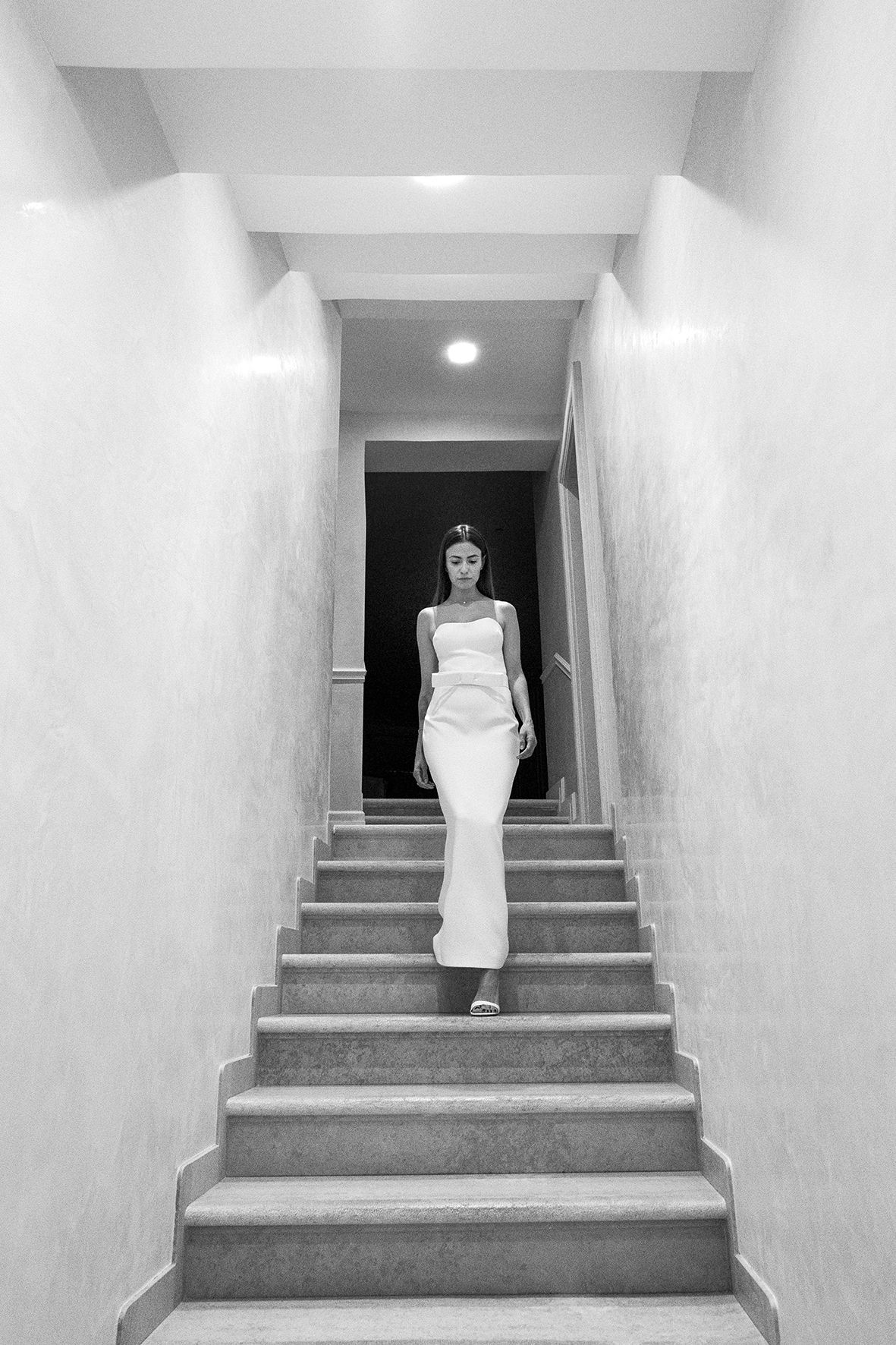 Black and white photo of a figure in a white formal gown standing on concrete stairs in a minimalist hallway.