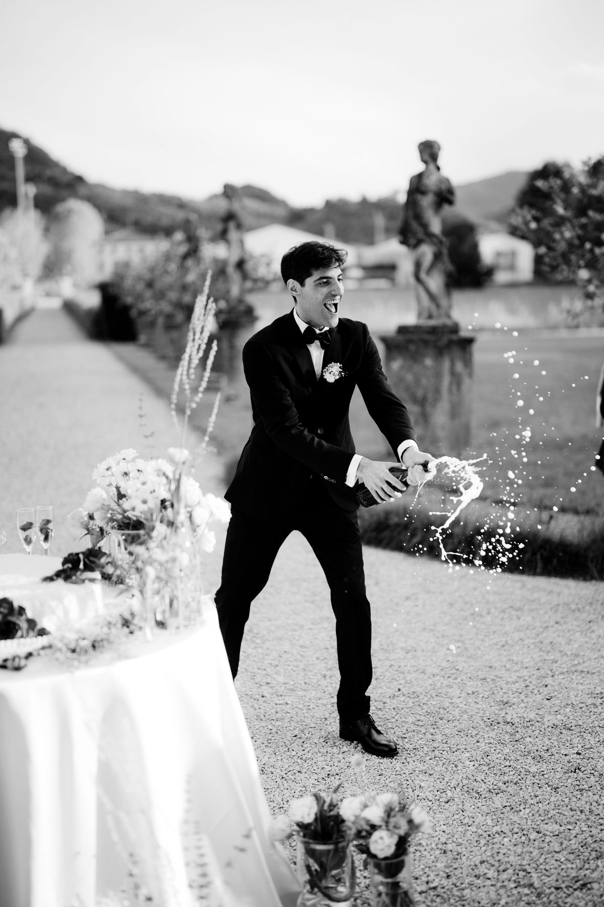 Groom in classic tuxedo playfully pops champagne in an elegant garden setting.