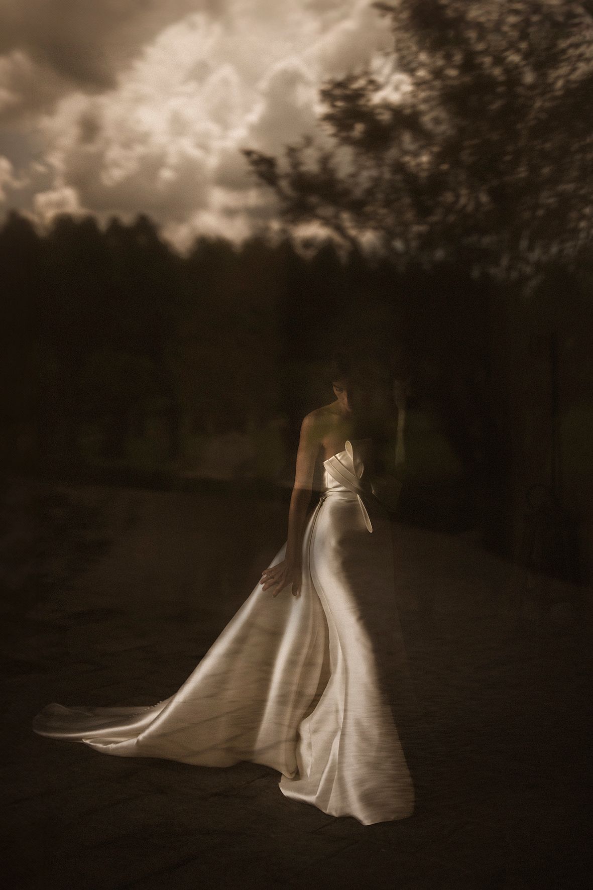 Sepia-toned photo of a wedding dress silhouette against a moody sky with trees in background.