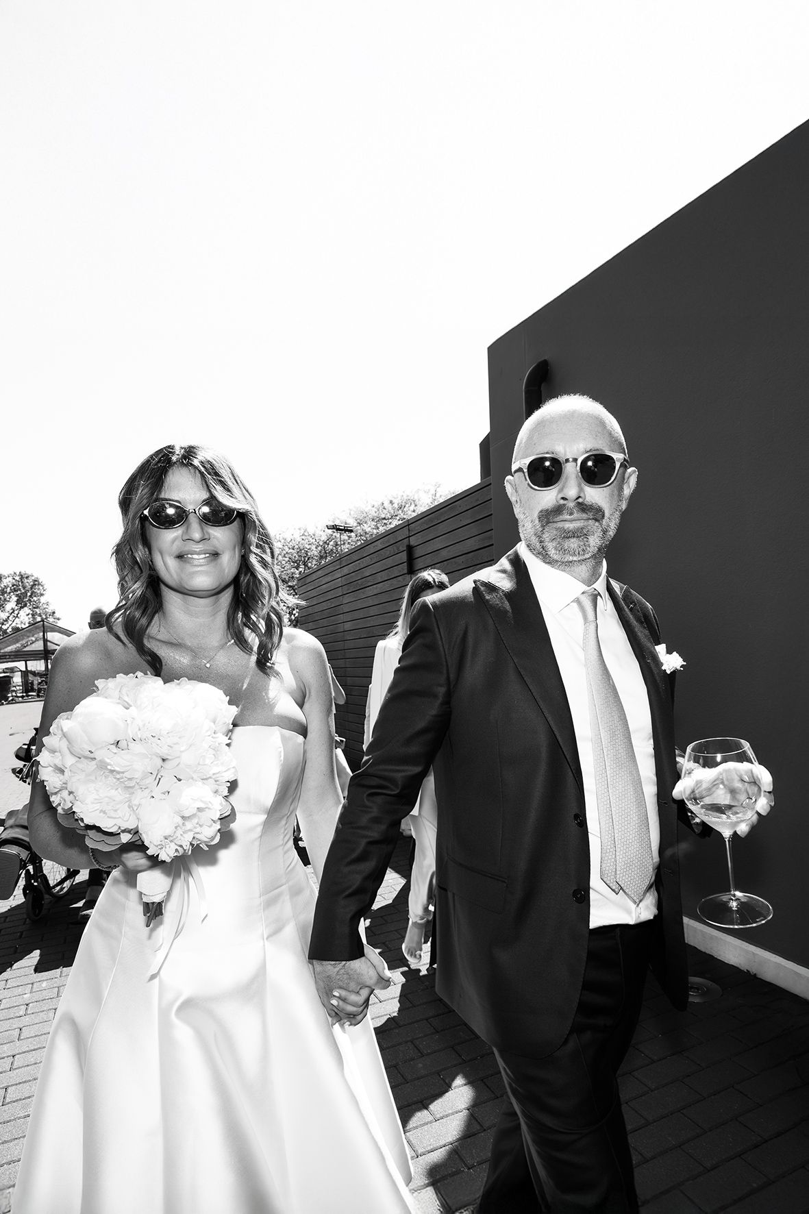 A black and white wedding photo showing the bride in a white dress with bouquet and companion in sunglasses on a deck.