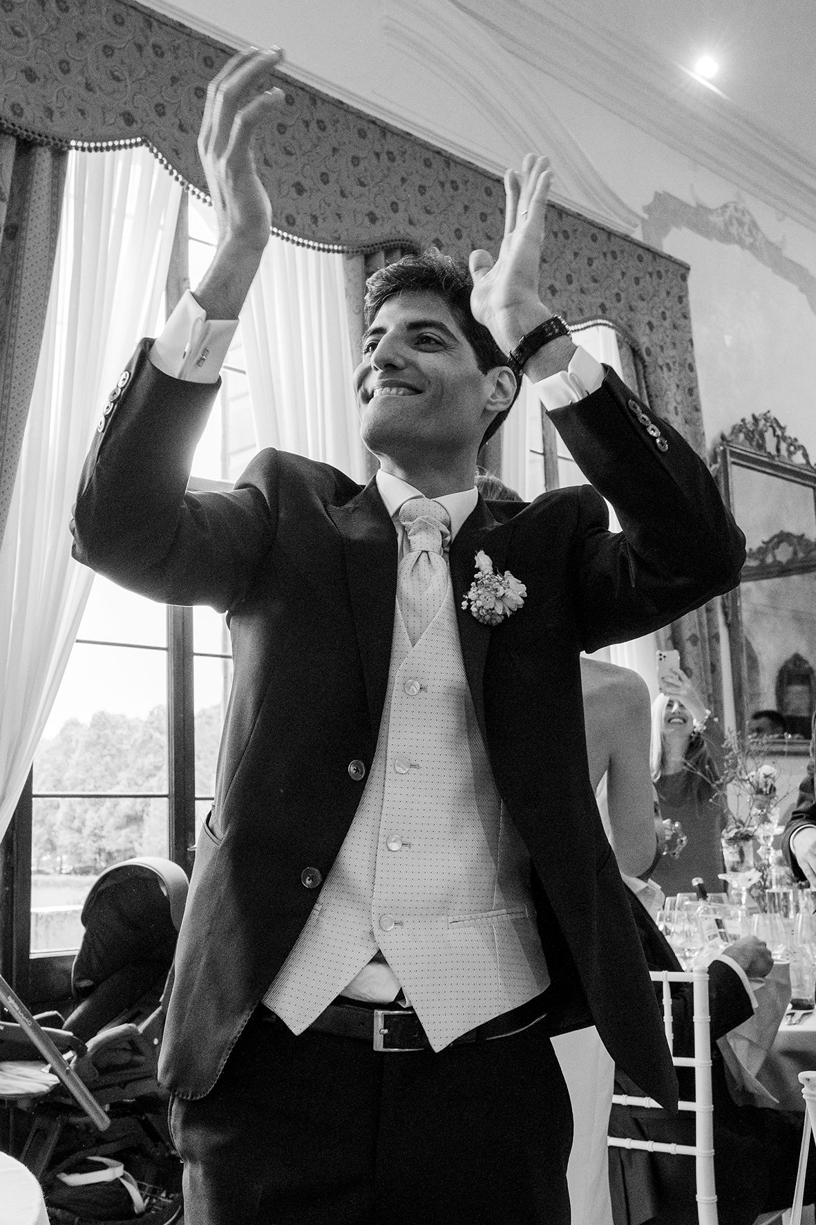 A joyful person in a formal black tuxedo and white vest raises their hands in celebration at an elegant indoor venue.