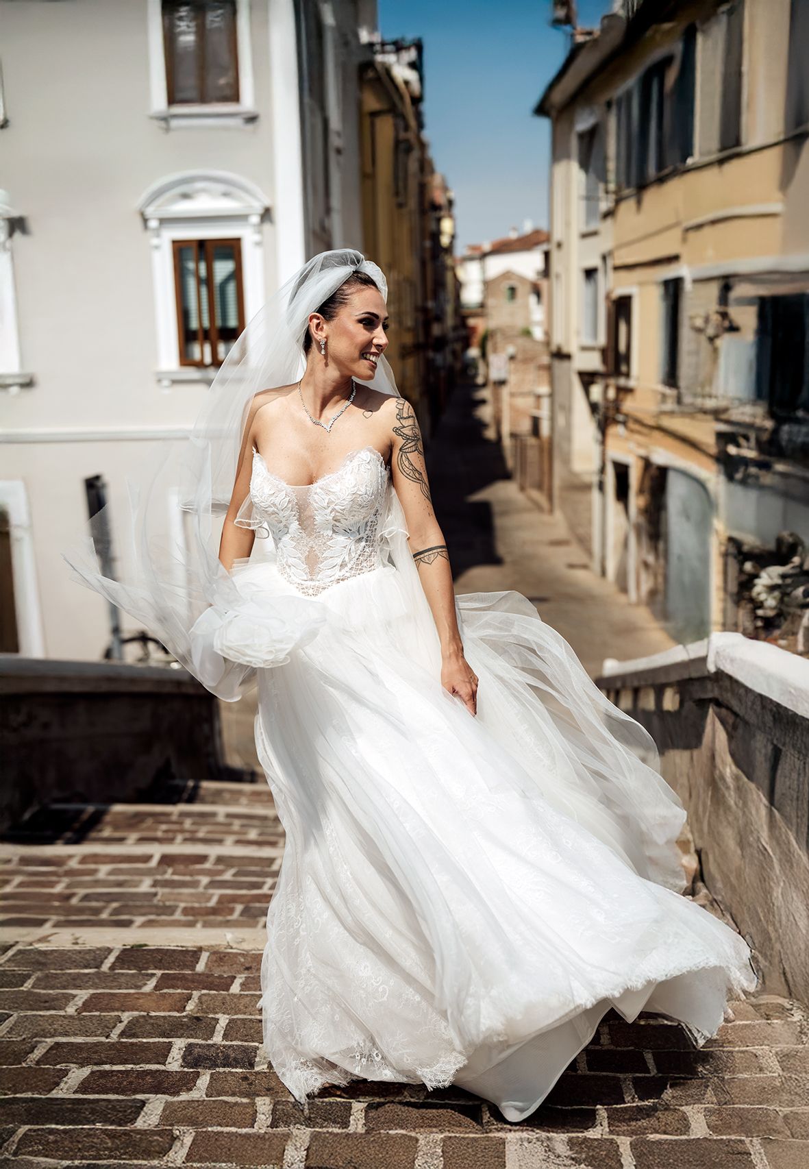 A bride in a flowing white lace gown stands on cobblestone steps in a charming Italian street.