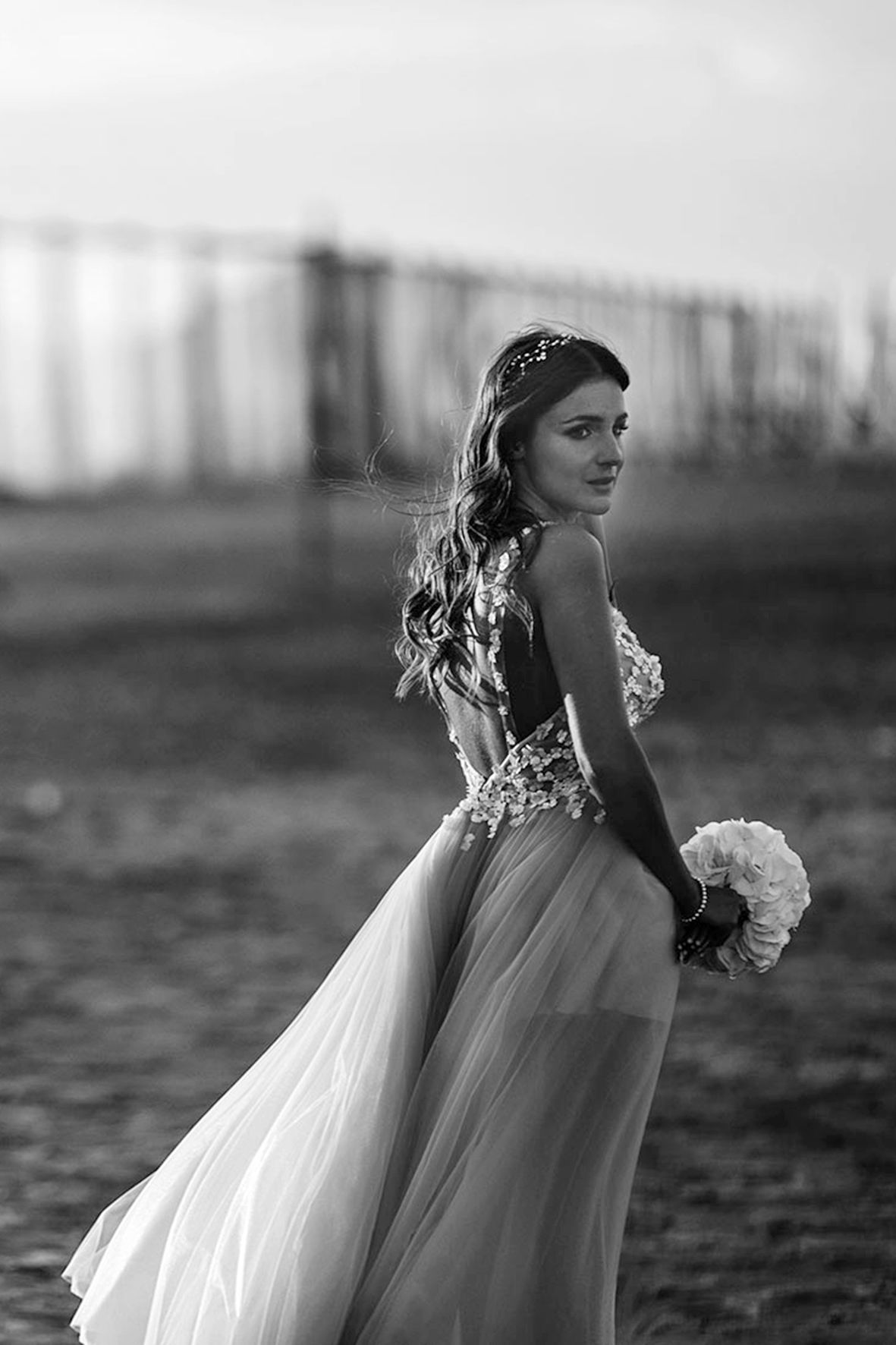 A bride in a flowing white gown with floral details poses outdoors at dusk while holding a bouquet in black and white.