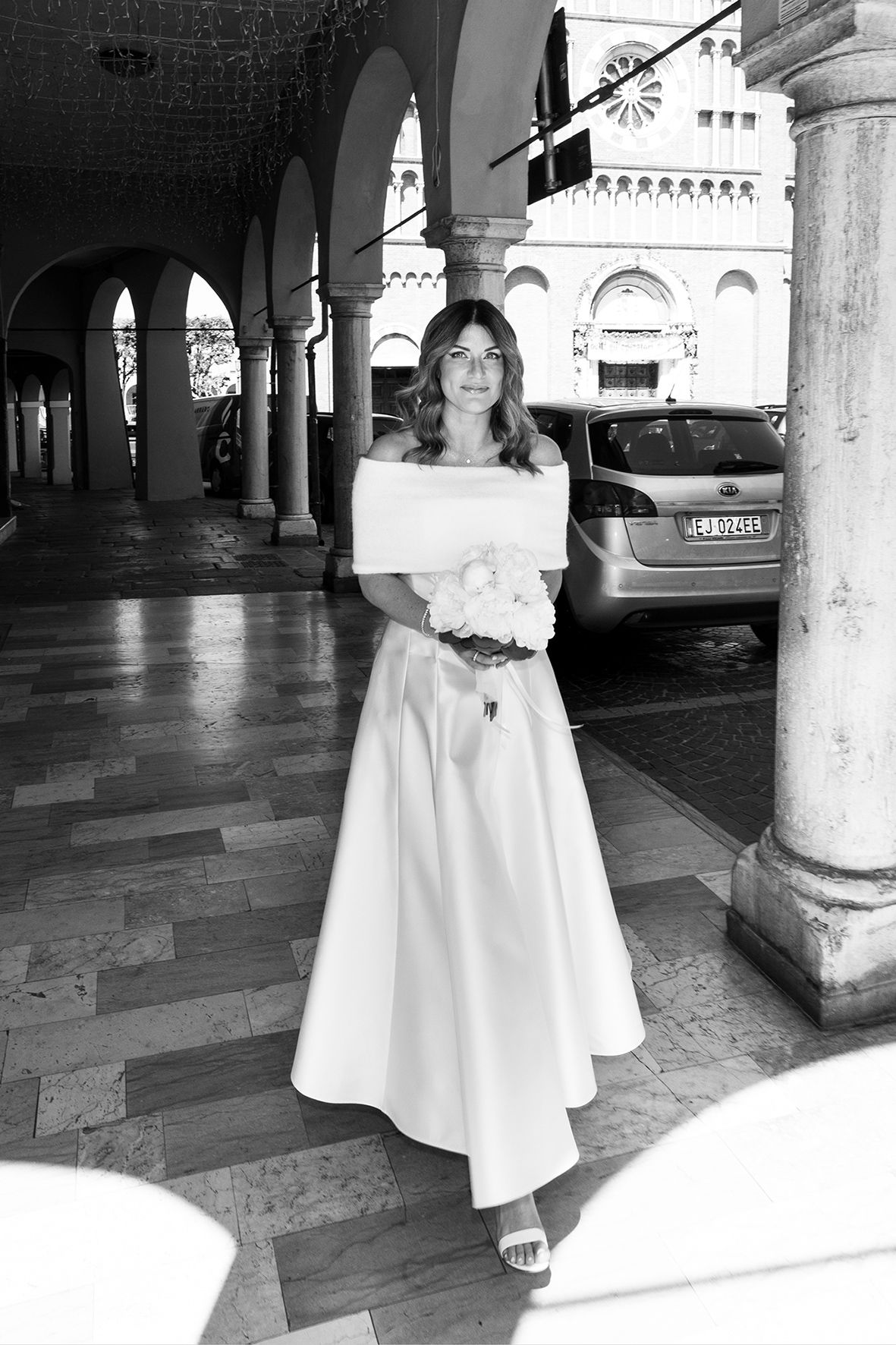 A bride in an off-shoulder white wedding gown stands under historic stone arches with a bouquet in black and white photography.