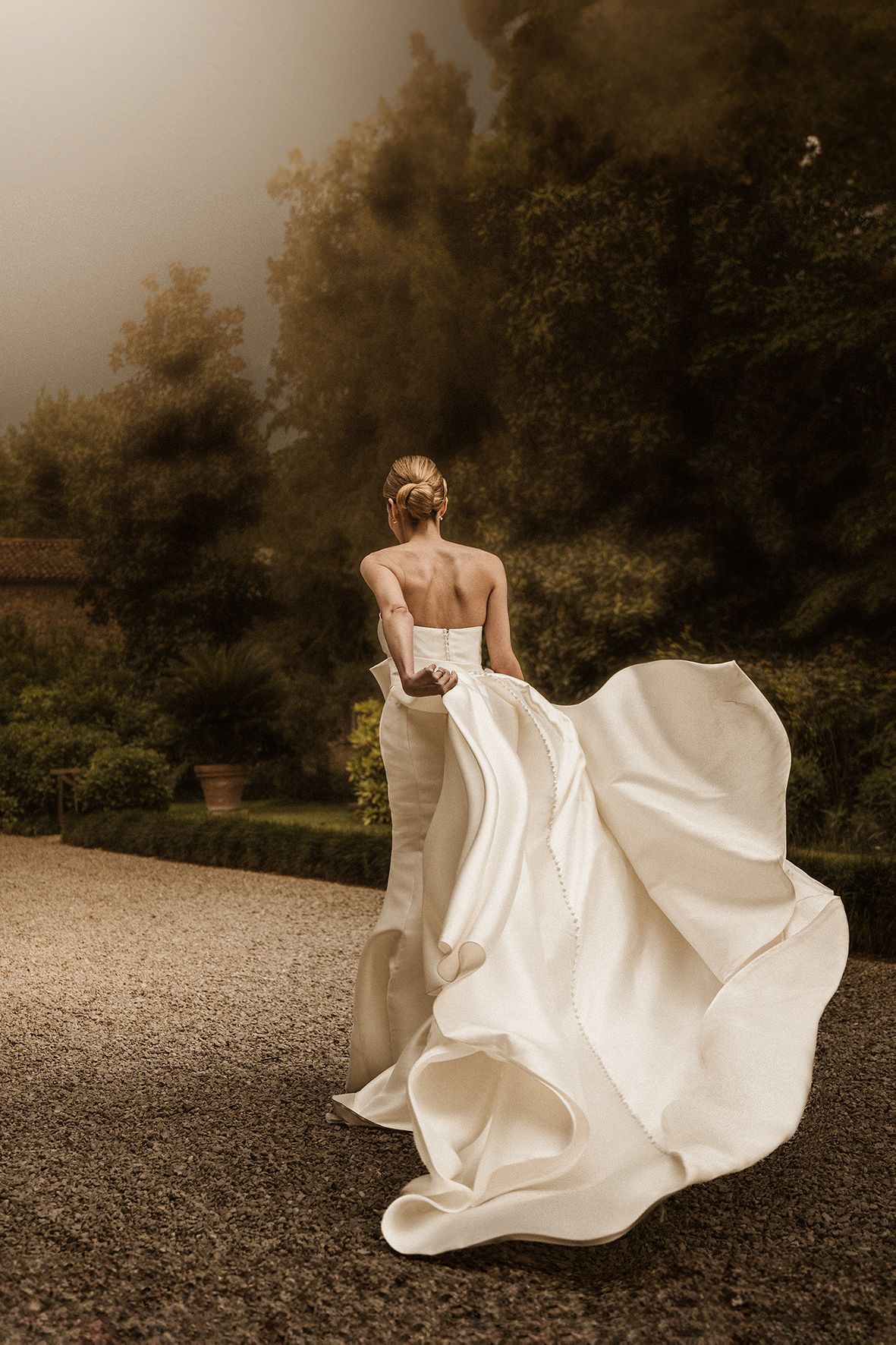 White wedding dress billows elegantly in the wind as bride twirls on gravel path with autumn trees in background.