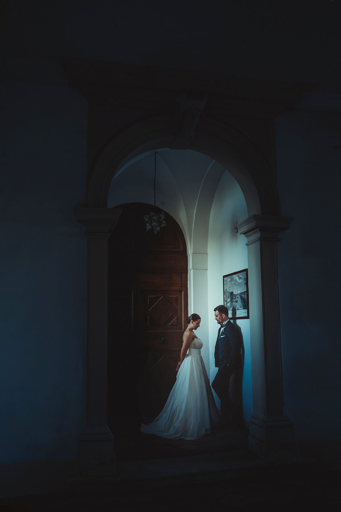 A couple in wedding attire stands intimately in a dimly lit archway with moody blue lighting creating a romantic atmosphere.
