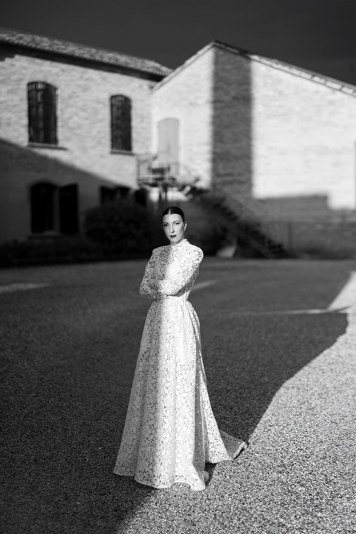 Black and white photo of a person in an elegant long dress standing on a path near a brick building in sunlight.