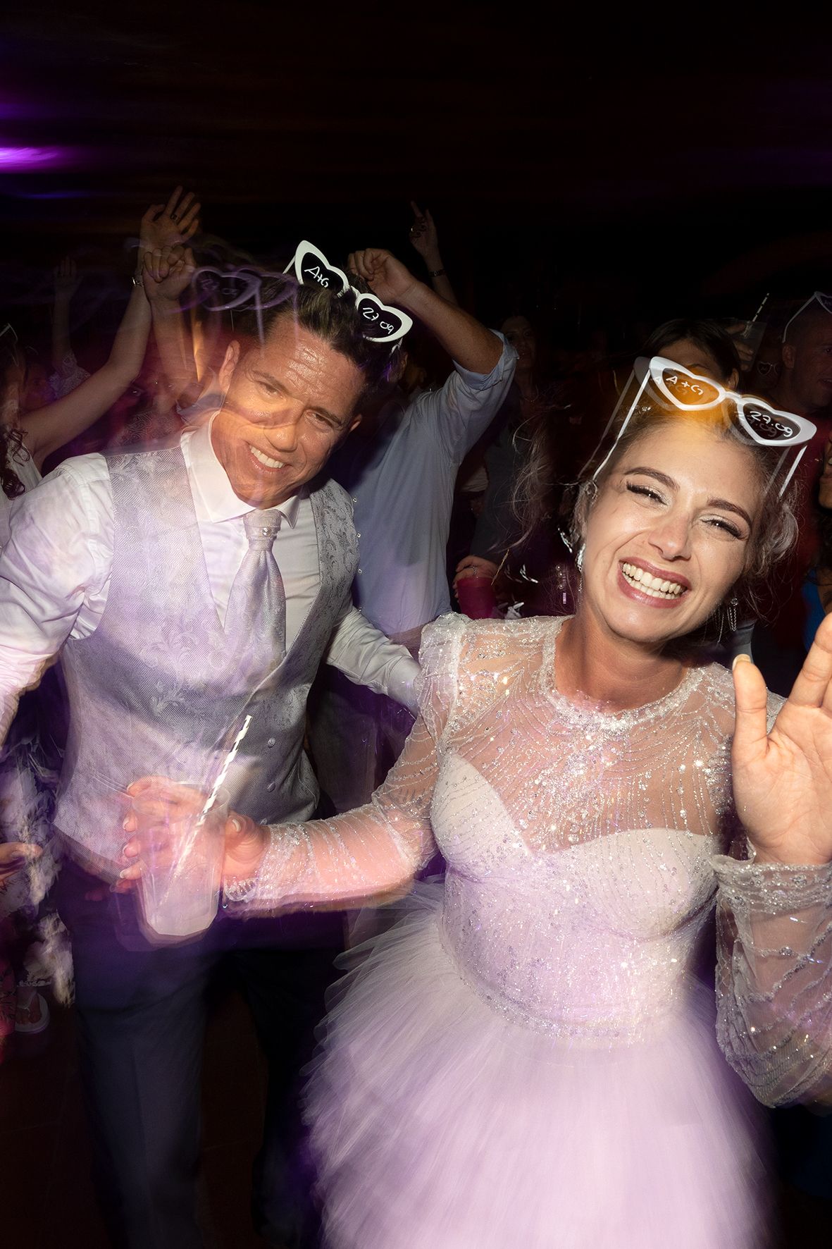 Party guests dancing and having fun on the dance floor wearing white outfits and novelty sunglasses at a celebration.