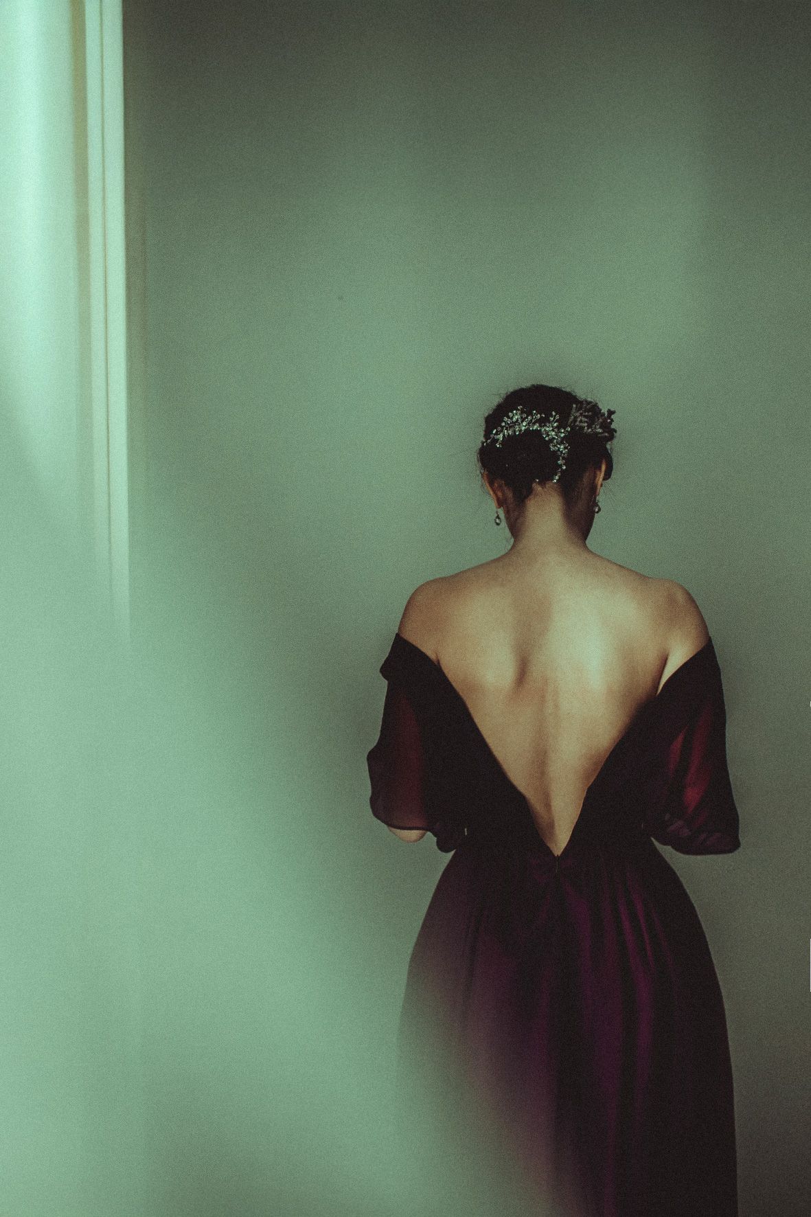 Artistic portrait showing bare back in deep burgundy dress against mint green wall with dramatic lighting and shadowing.