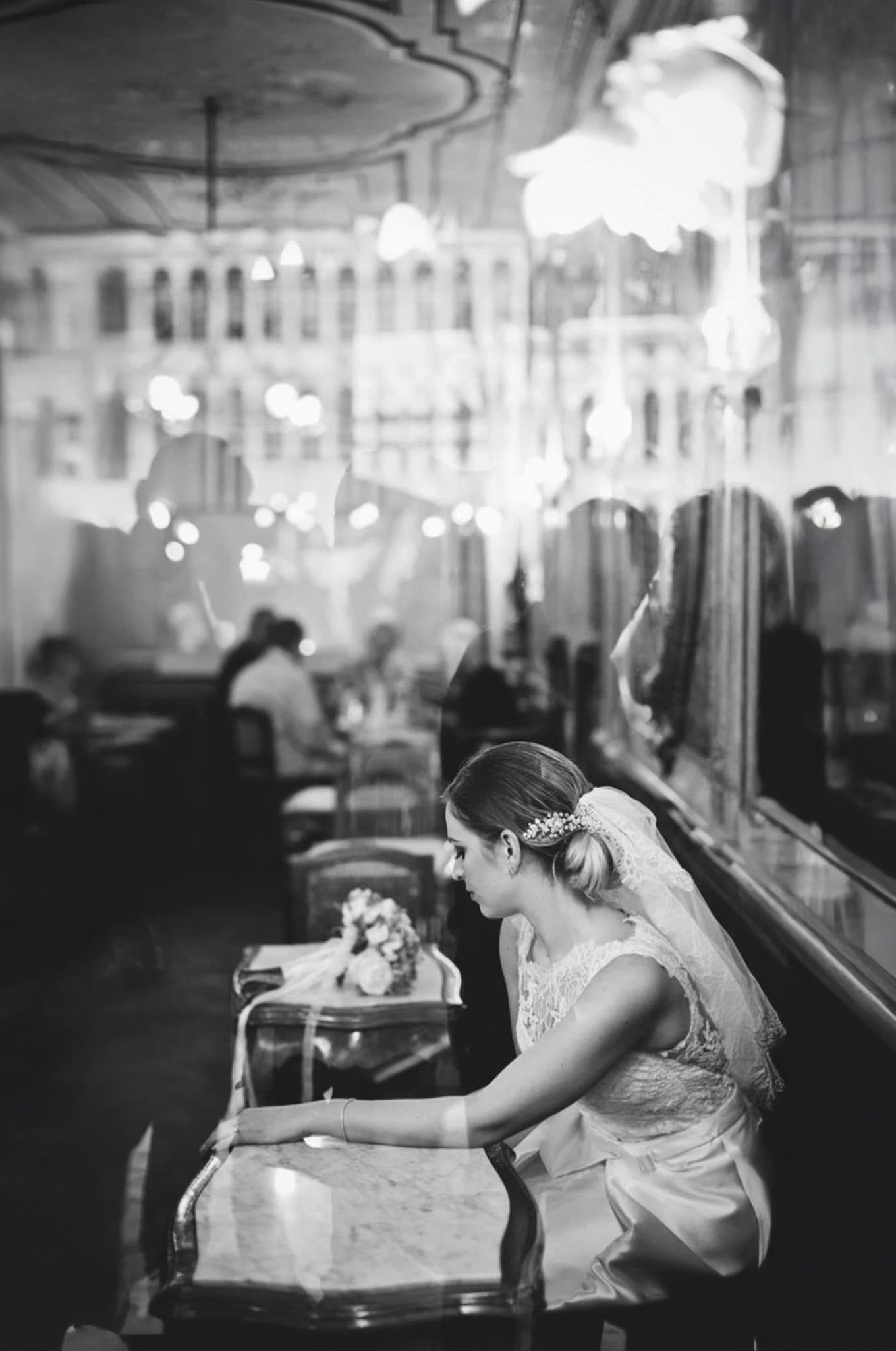 Bride in vintage-inspired dress sits at ornate bar under crystal chandeliers.