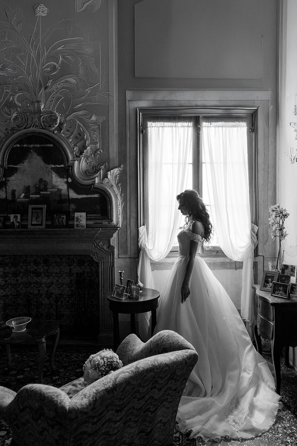 Black and white image of figure in flowing white wedding gown standing near window in vintage-style room with ornate decor.