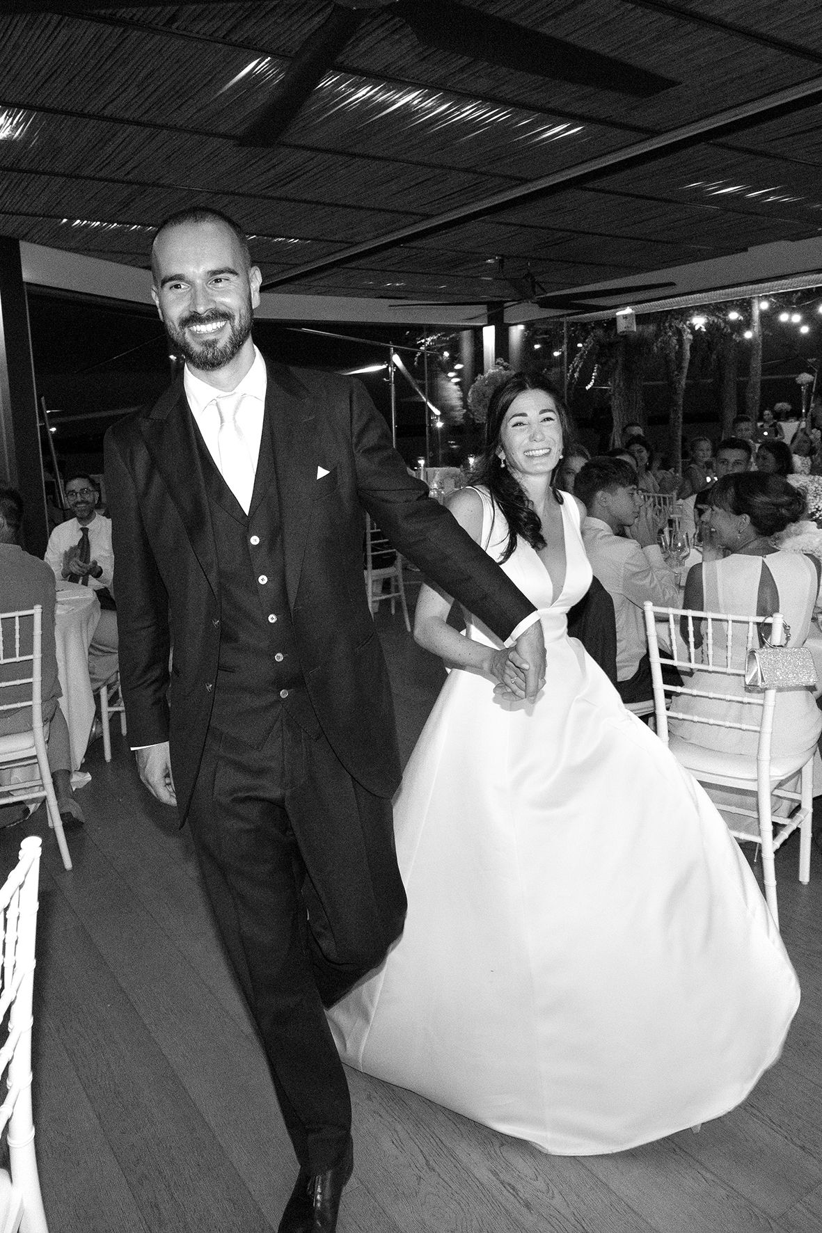 Joyful wedding couple laughs and dances at their reception venue with white chairs and evening lighting.