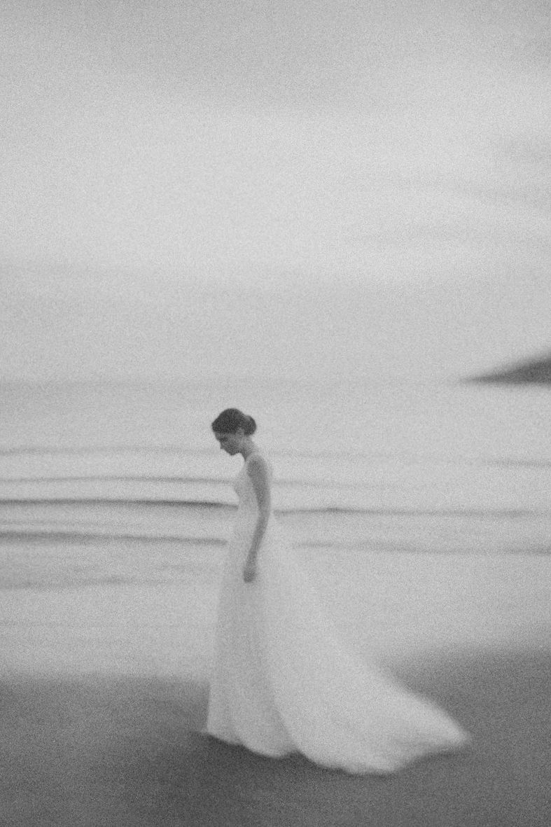 A dreamy black and white photo of a figure in a flowing white wedding gown standing alone on a misty beach.