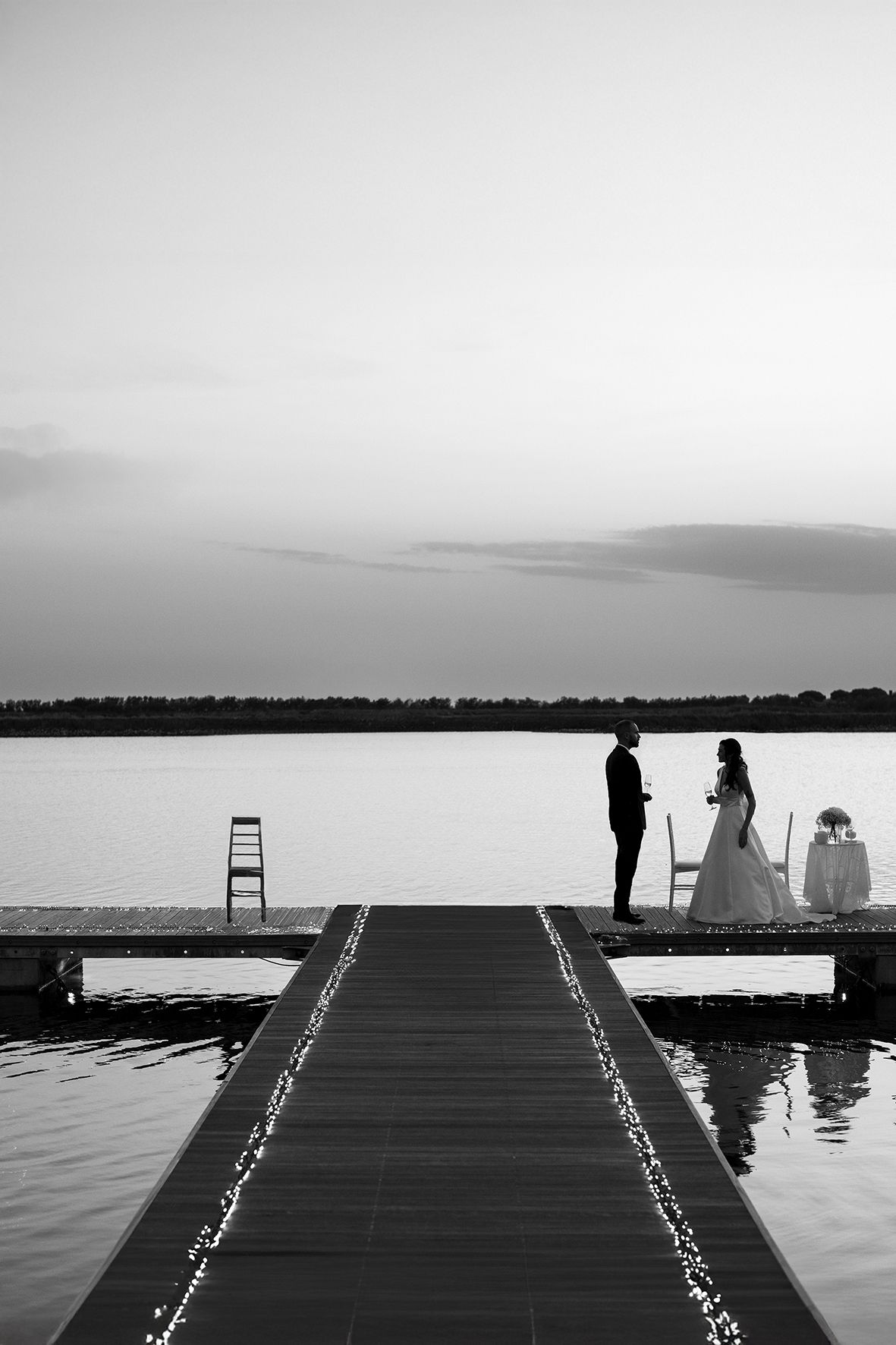 A silhouetted couple stands on a dock decorated with string lights at sunset, creating a romantic lakeside scene in black and white.