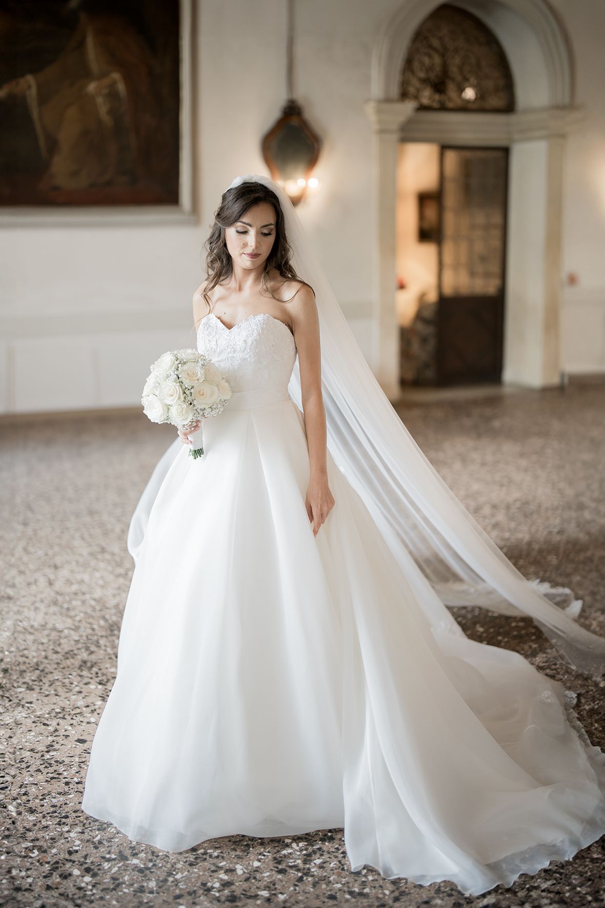 A bride in an elegant white strapless ballgown with long veil holds a white bouquet in an ornate historic interior.