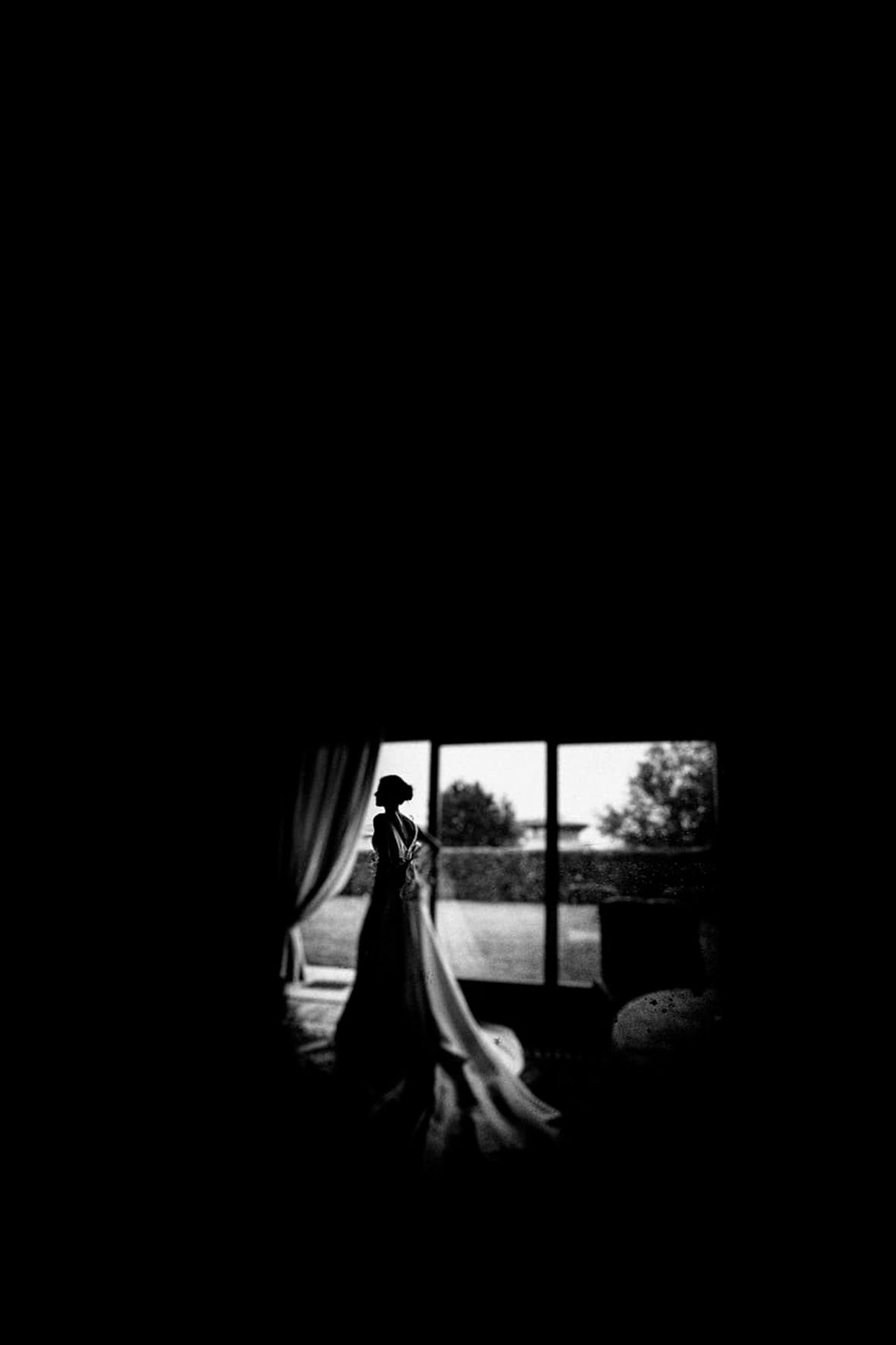 Silhouette of a bride by a window in black and white with dramatic lighting.