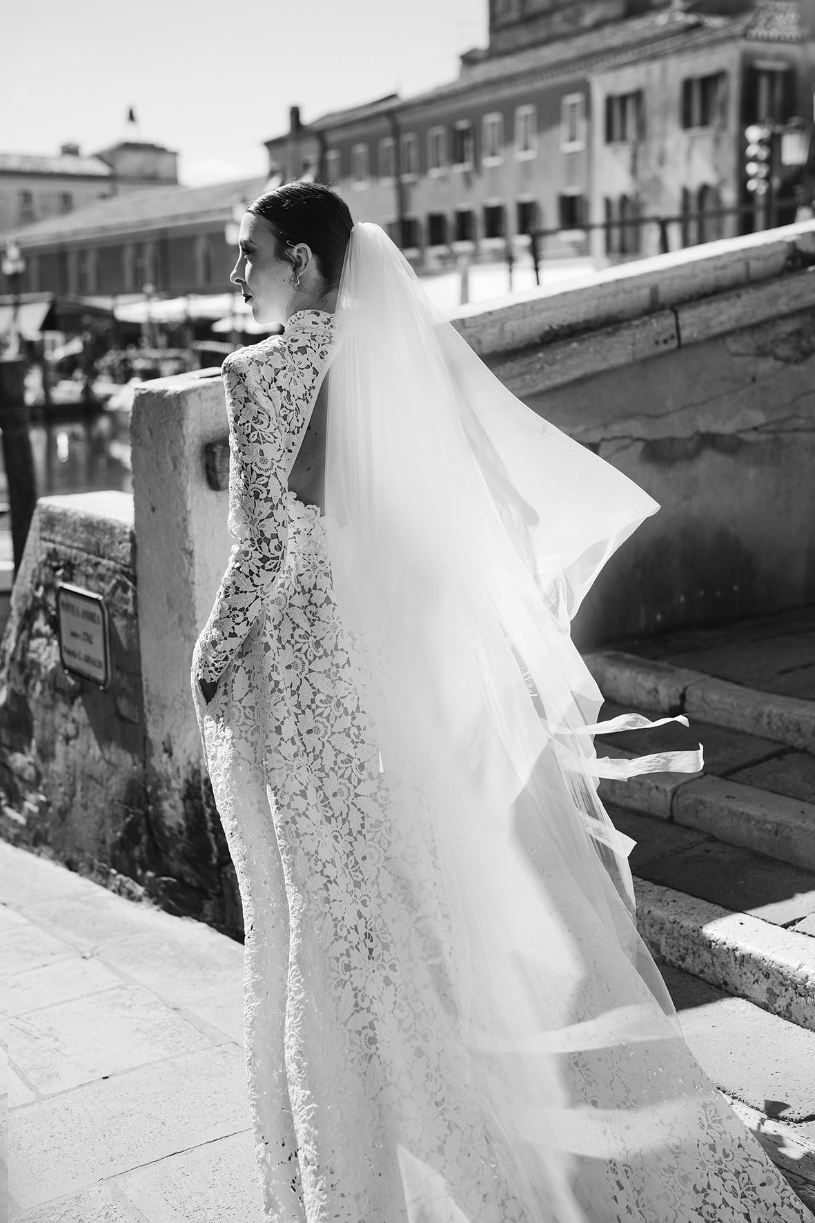 A bride in a fitted lace wedding gown and long flowing veil stands on stone steps in a black and white photograph.