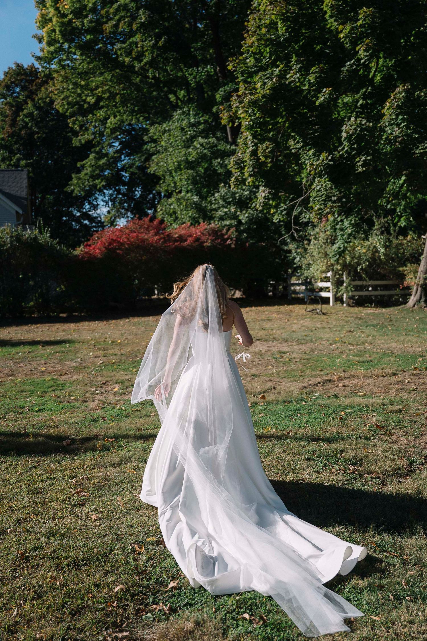 A Covet Bridal bride in a flowing white wedding dress and veil walks across a sunlit lawn with autumn trees in the background.
