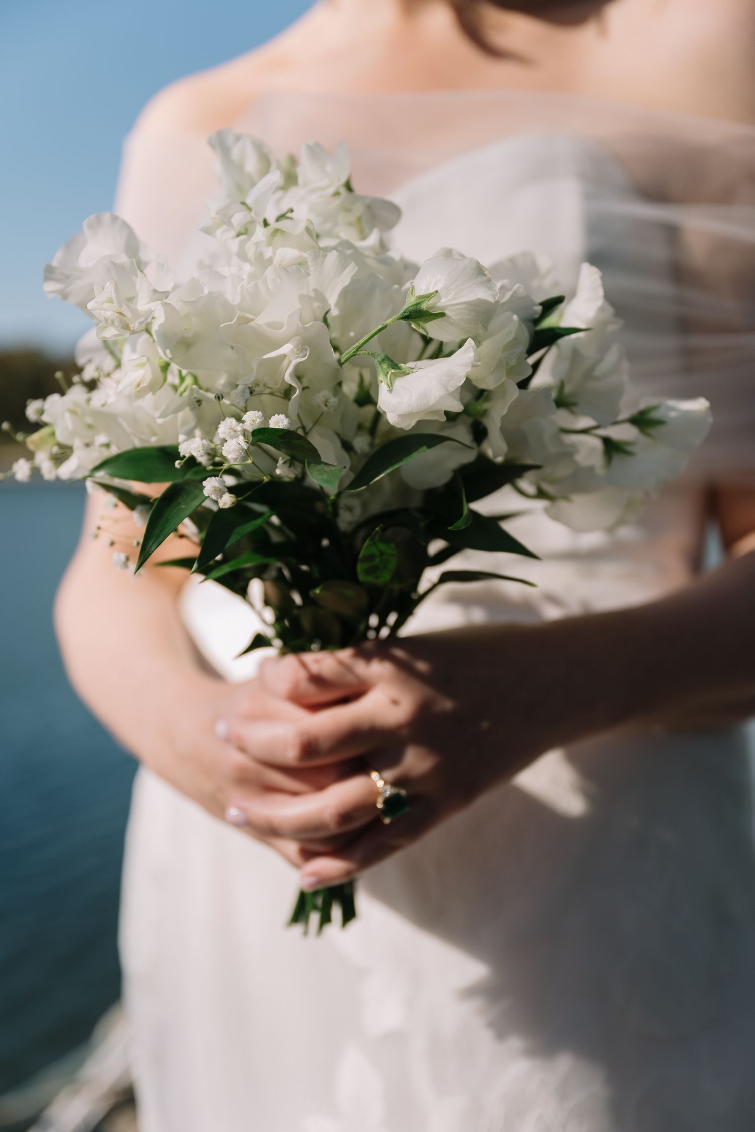 A delicate white bridal bouquet with lush flowers and green foliage held against a white wedding dress near water.